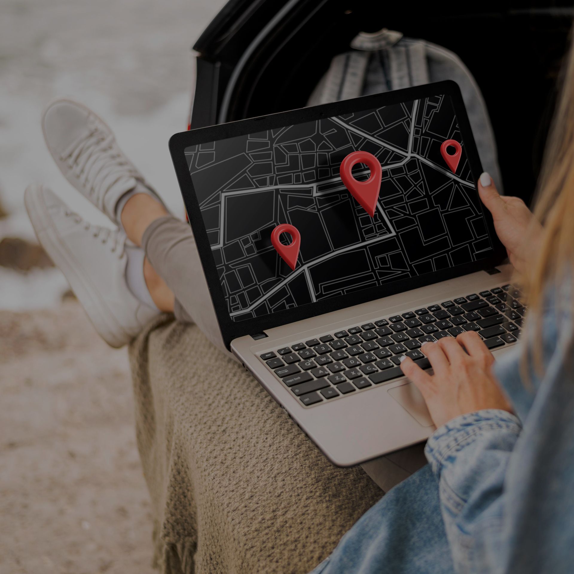 Person using a laptop with map and red location markers, sitting in a car trunk.