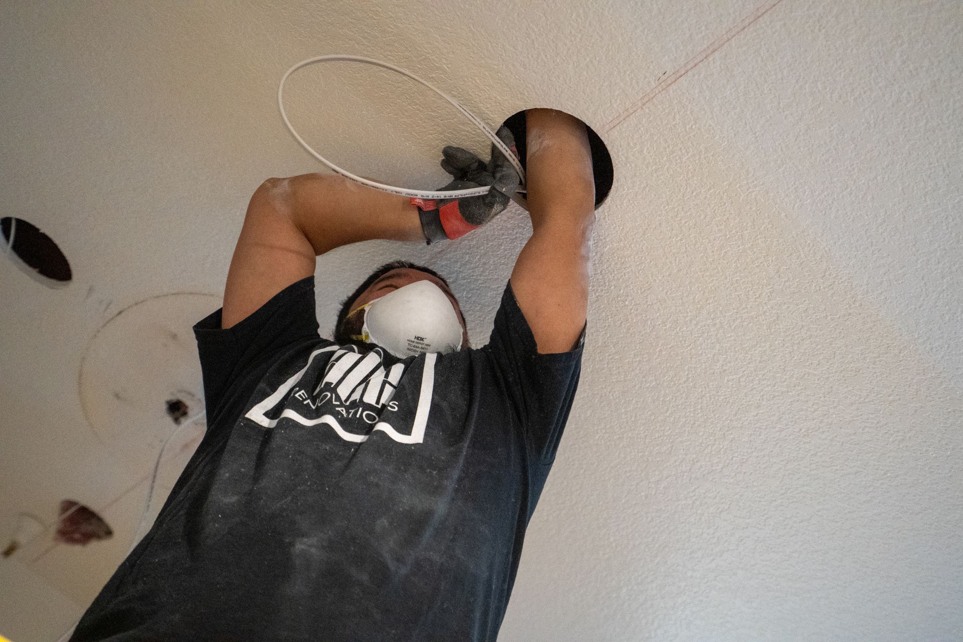 Person hanging a white cable from a ceiling in a room, wearing a black shirt and white face mask