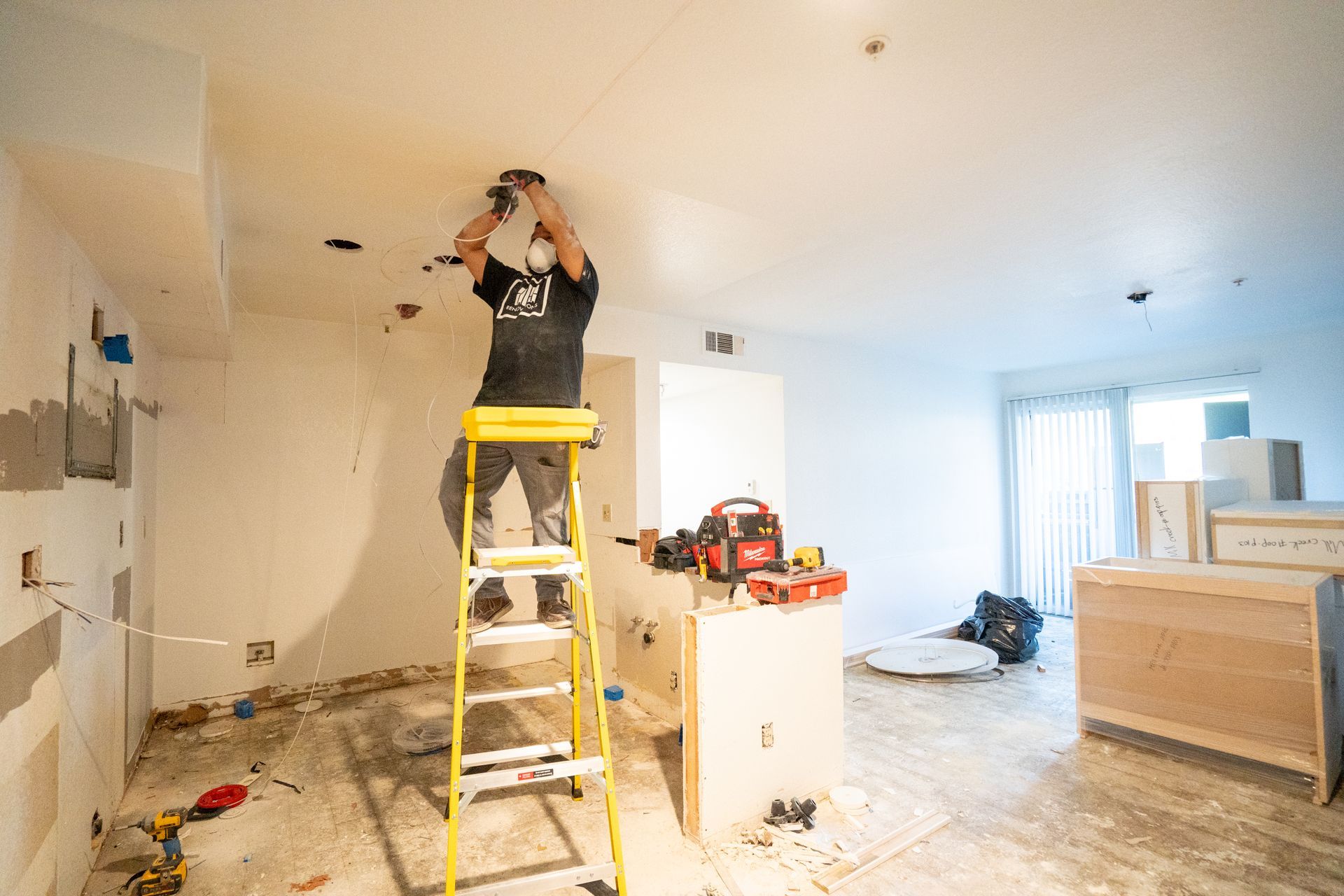 Worker on a yellow ladder installing a ceiling fixture in a bright, unfinished room