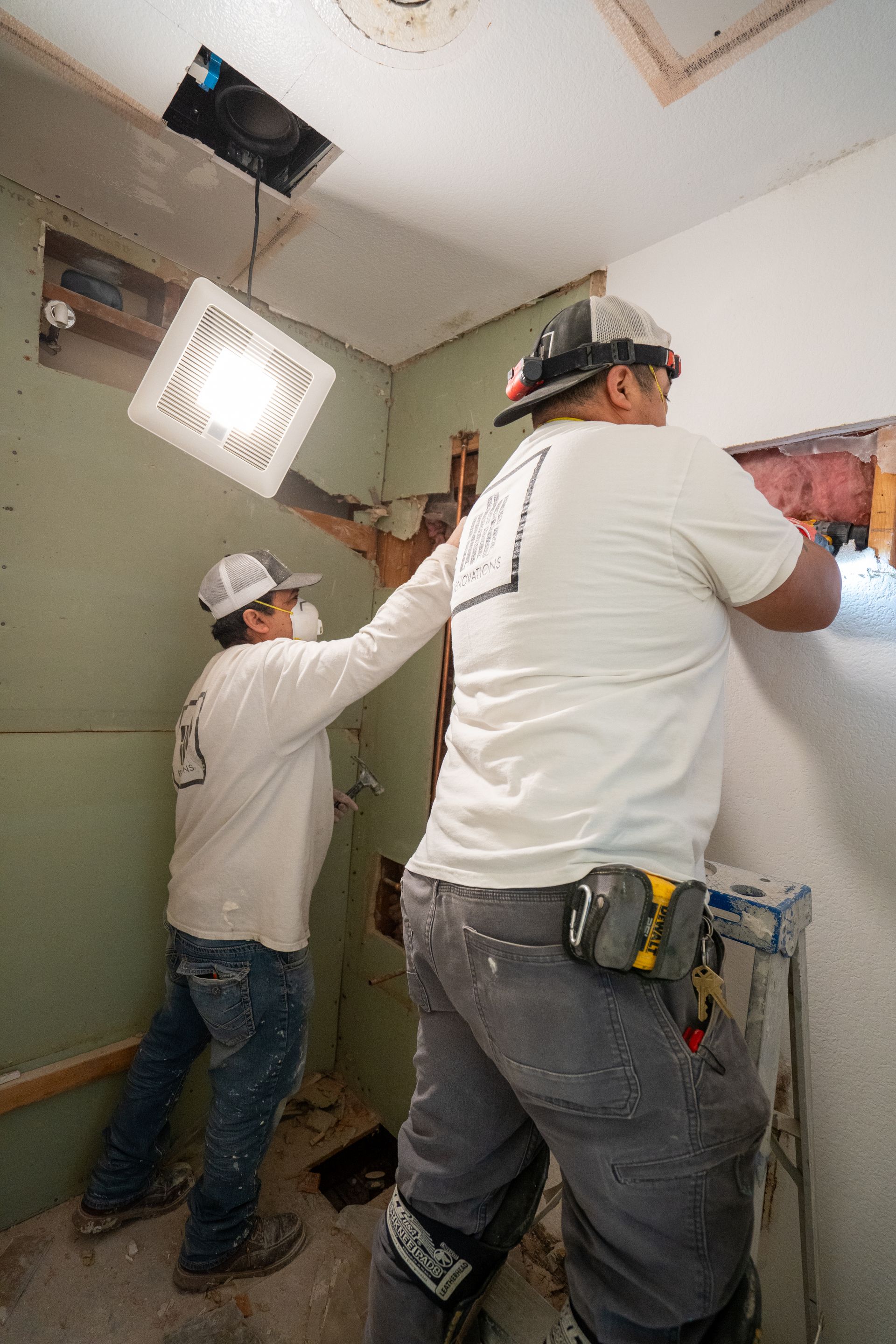Two workers repairing a ceiling with tools in a room, one on a ladder under a bright work light