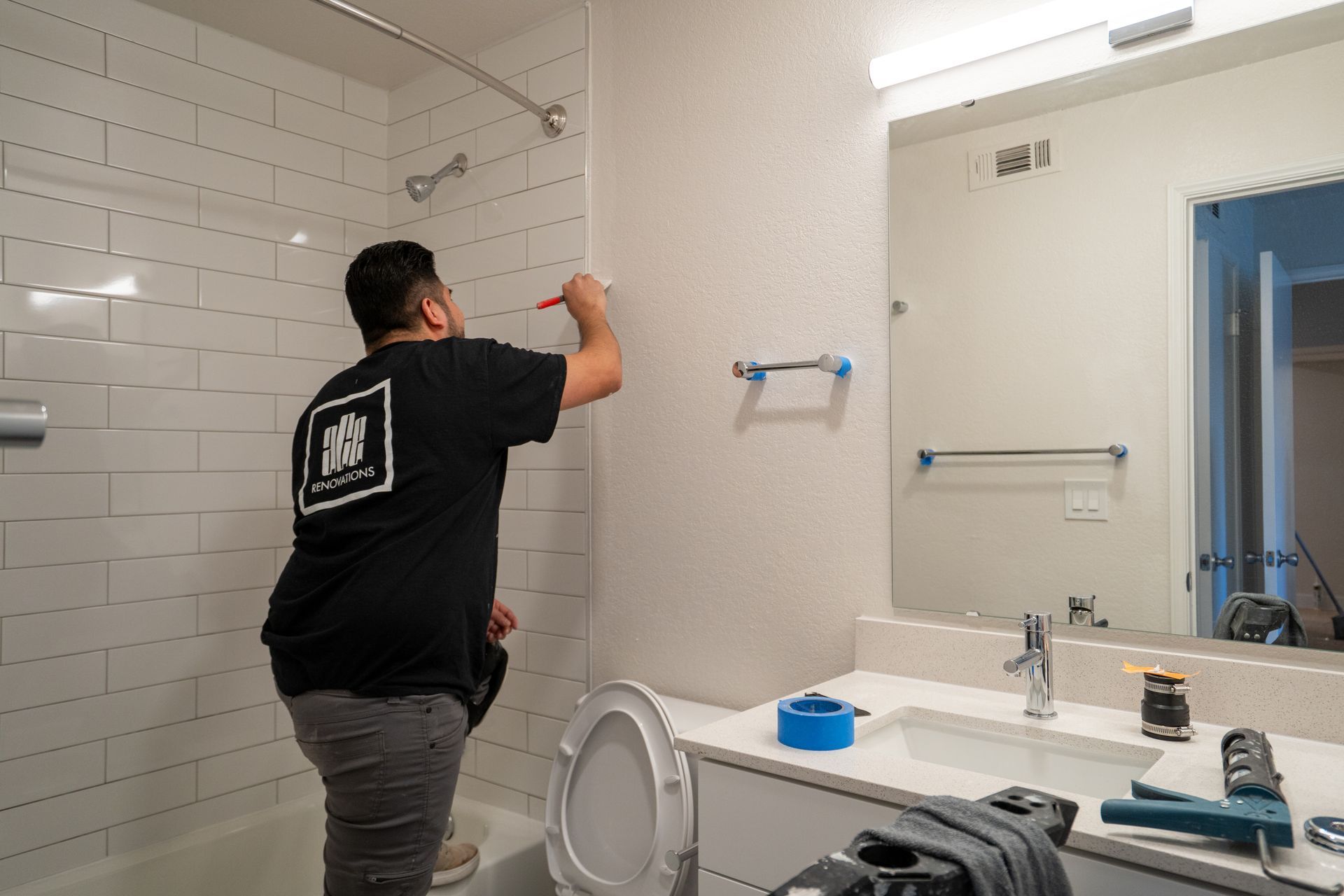 Person painting a bathroom wall beside a mirror and sink with blue painter’s tape.