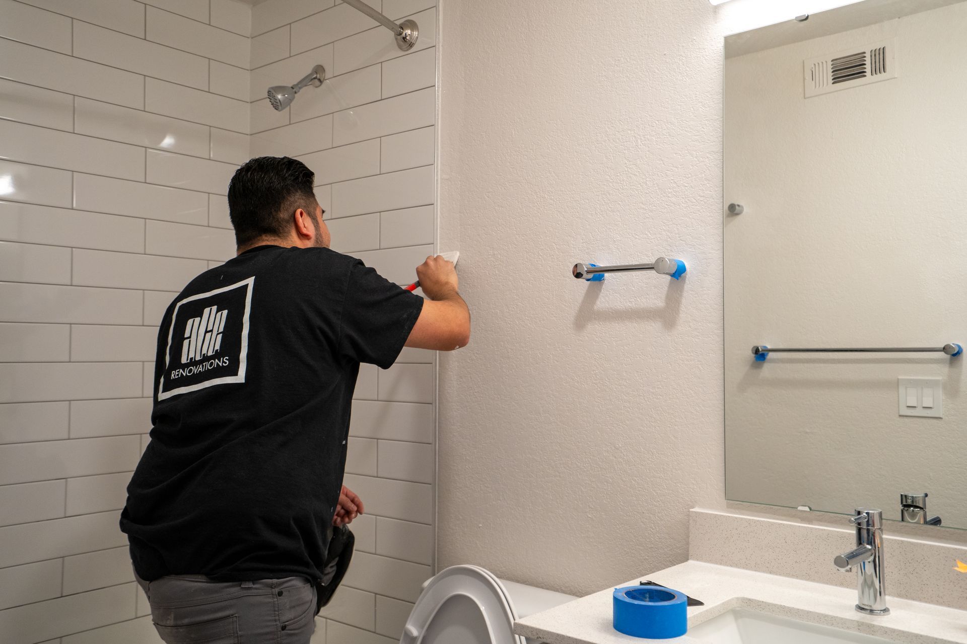 Man installing bathroom mirror strip with tape, next to a sink and toilet in a tiled bathroom