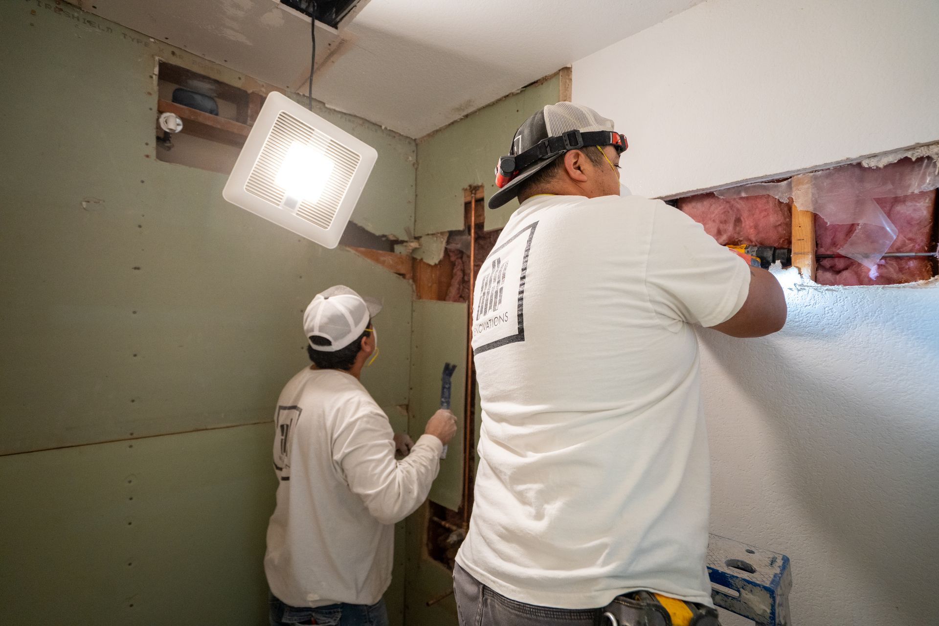 Two workers installing wall tile in a bathroom, one standing on a ladder under bright work lights.
