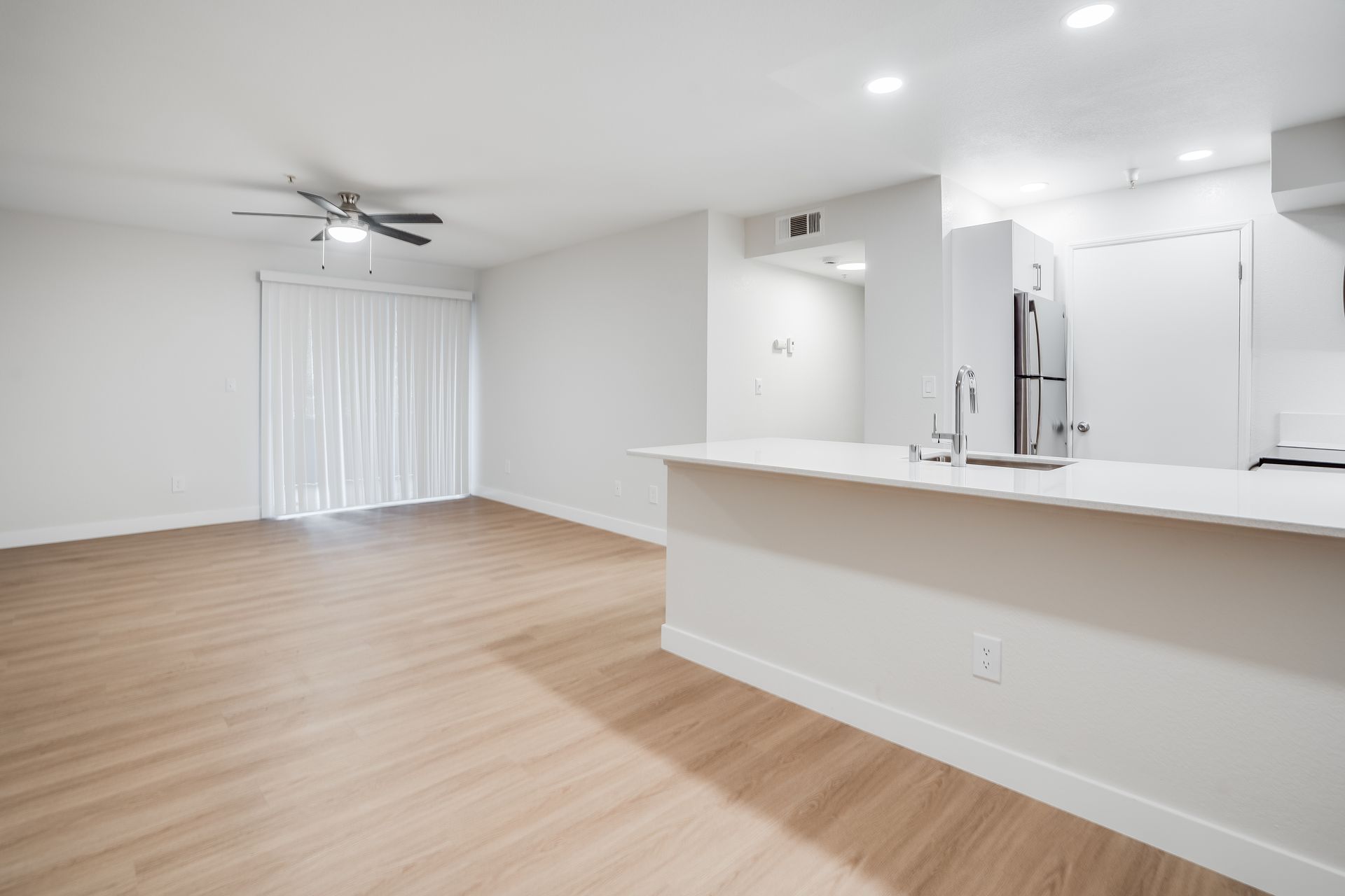Bright empty open-plan living room with light wood floors, white walls, ceiling fan, and kitchen bar counter