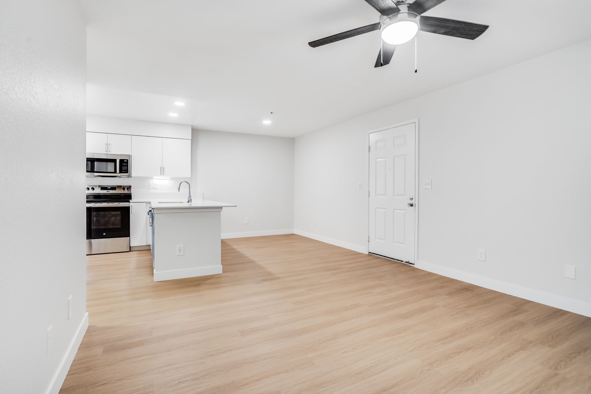 Bright empty living room with light wood floors, white walls, ceiling fan, and small kitchen area.