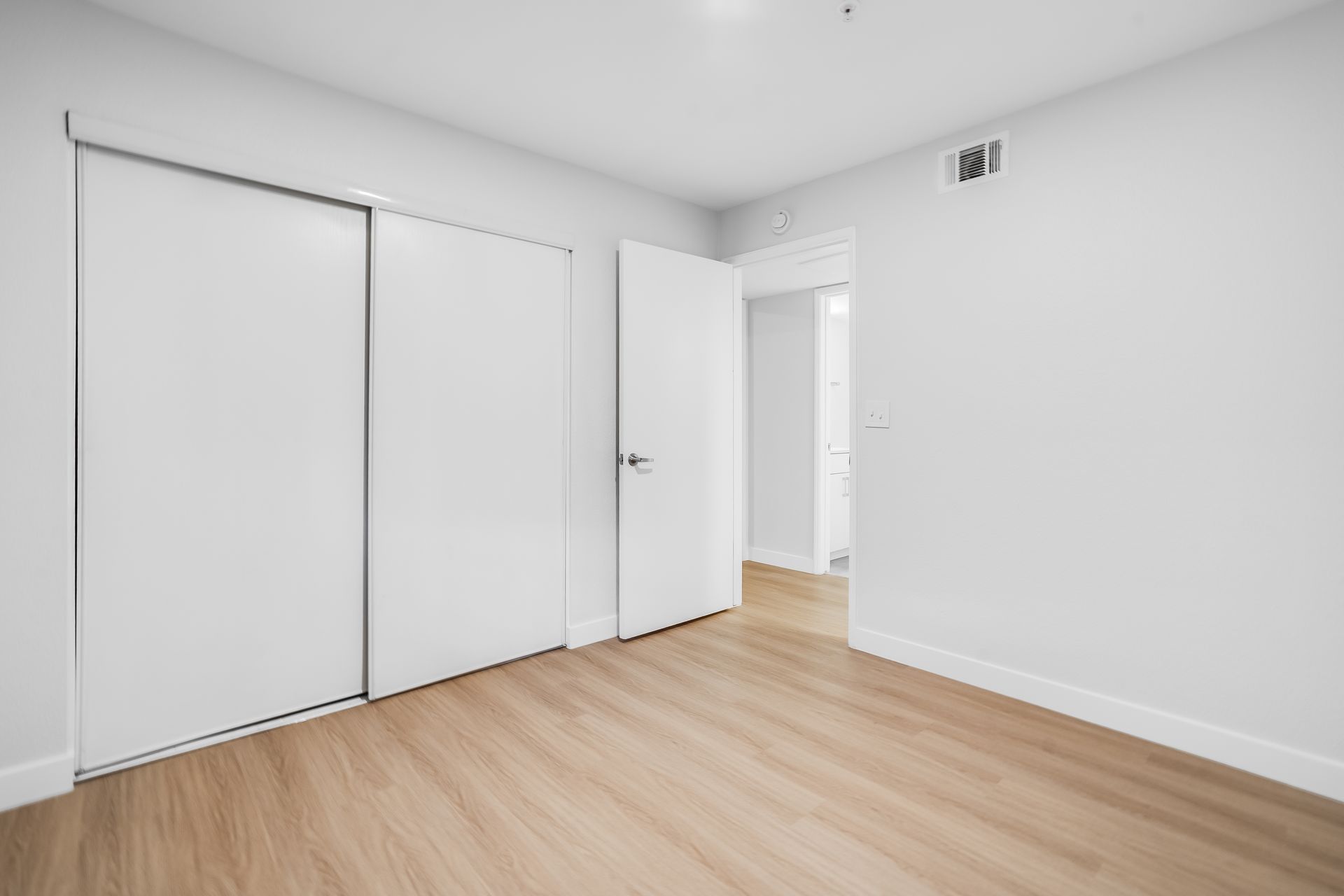 Empty white bedroom with wood flooring, open closet doors, and a doorway to a hall
