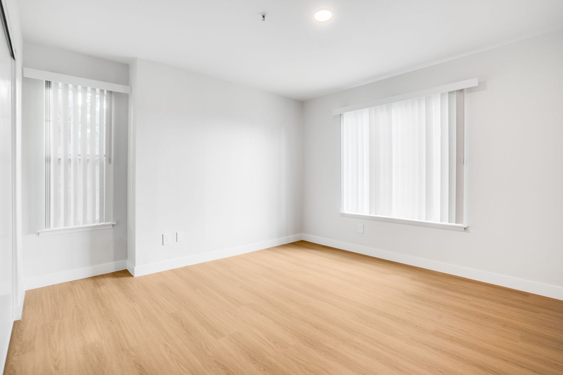 Empty bright room with light wood floors, white walls, and windows with blinds.