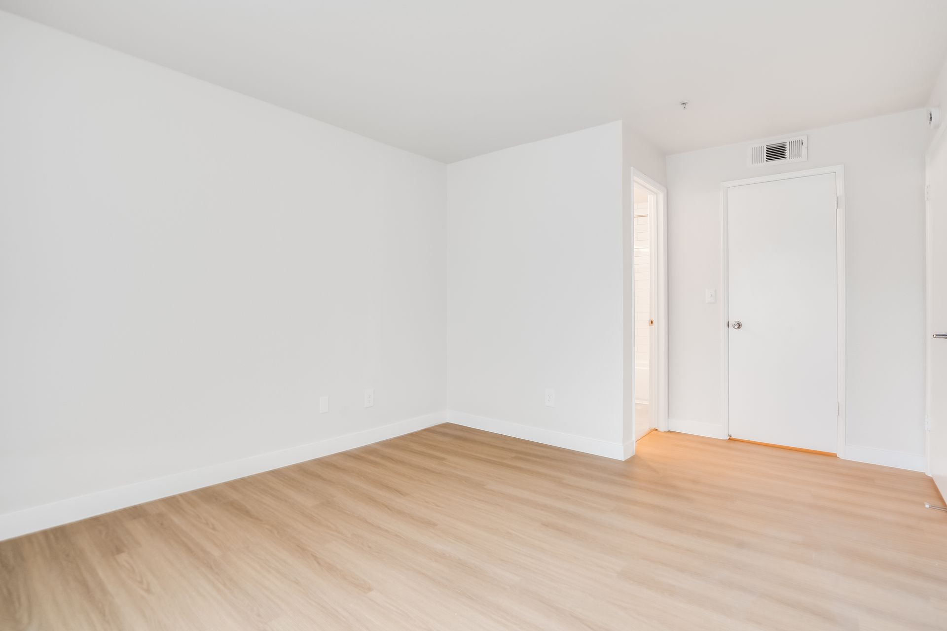 Empty white room with light wood floors and a closed door near a small entryway.
