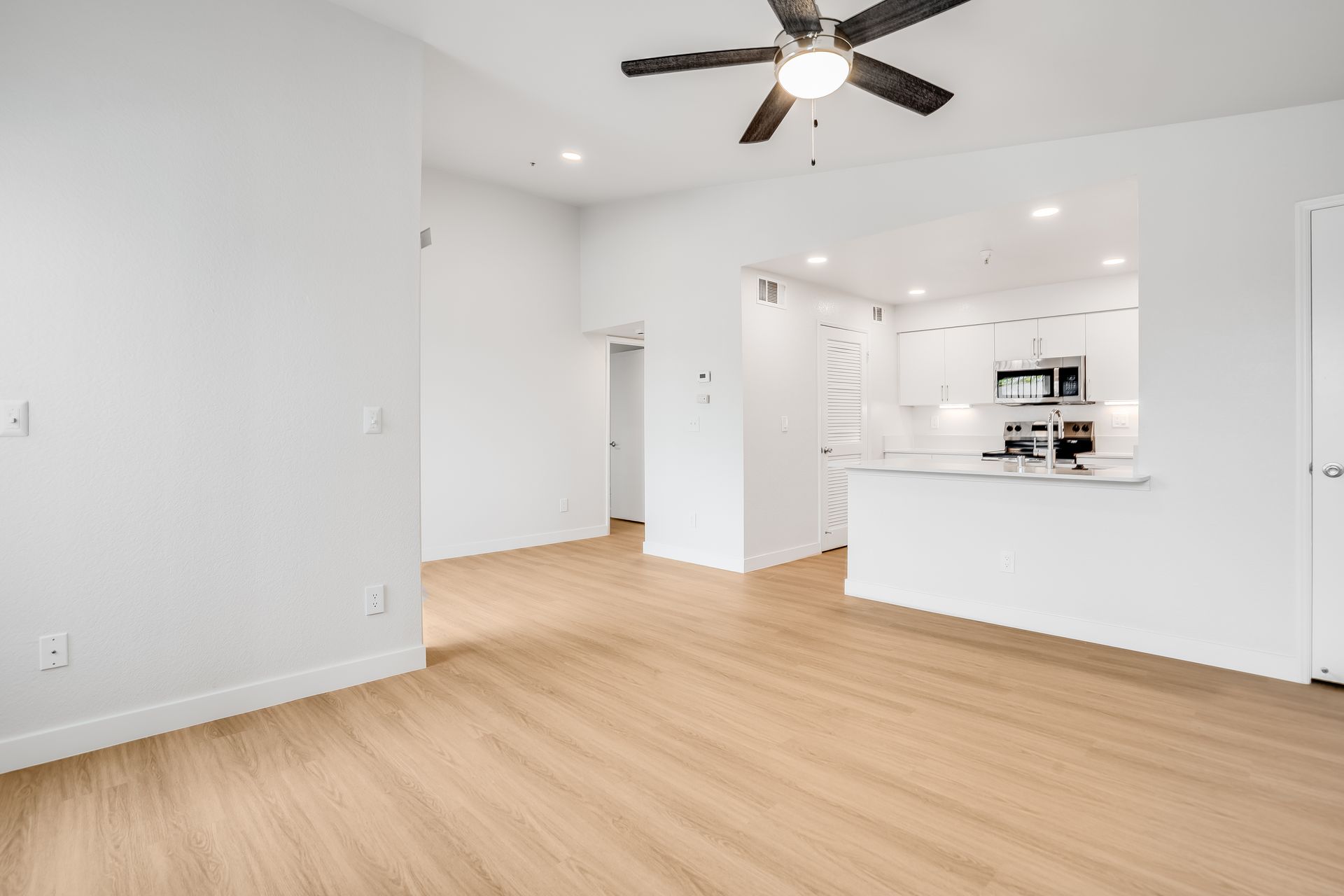 Empty modern living room with light wood floors, white walls, and an open kitchen under a ceiling fan