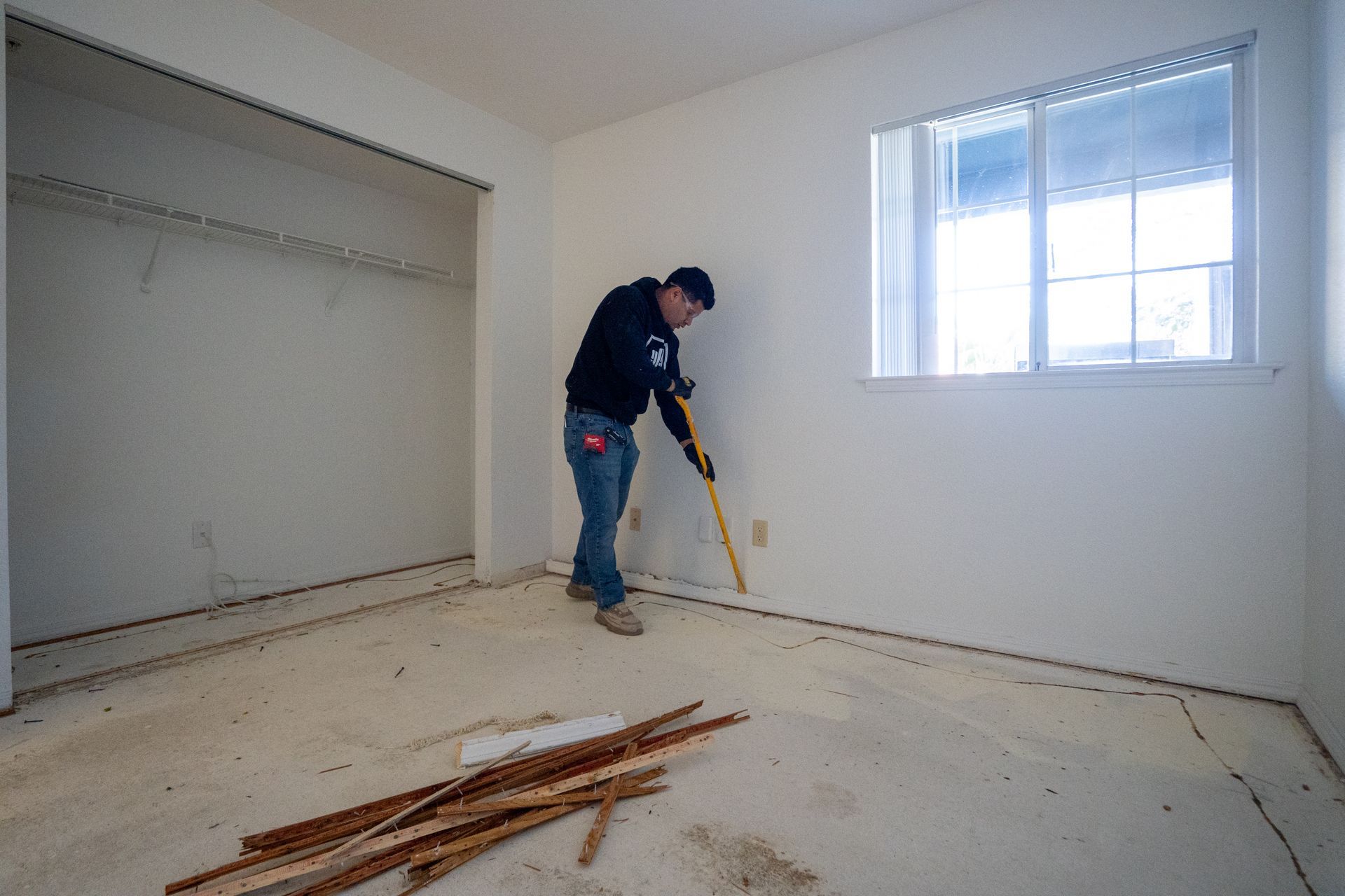 Person sweeping debris in a mostly empty room with white walls and a window.