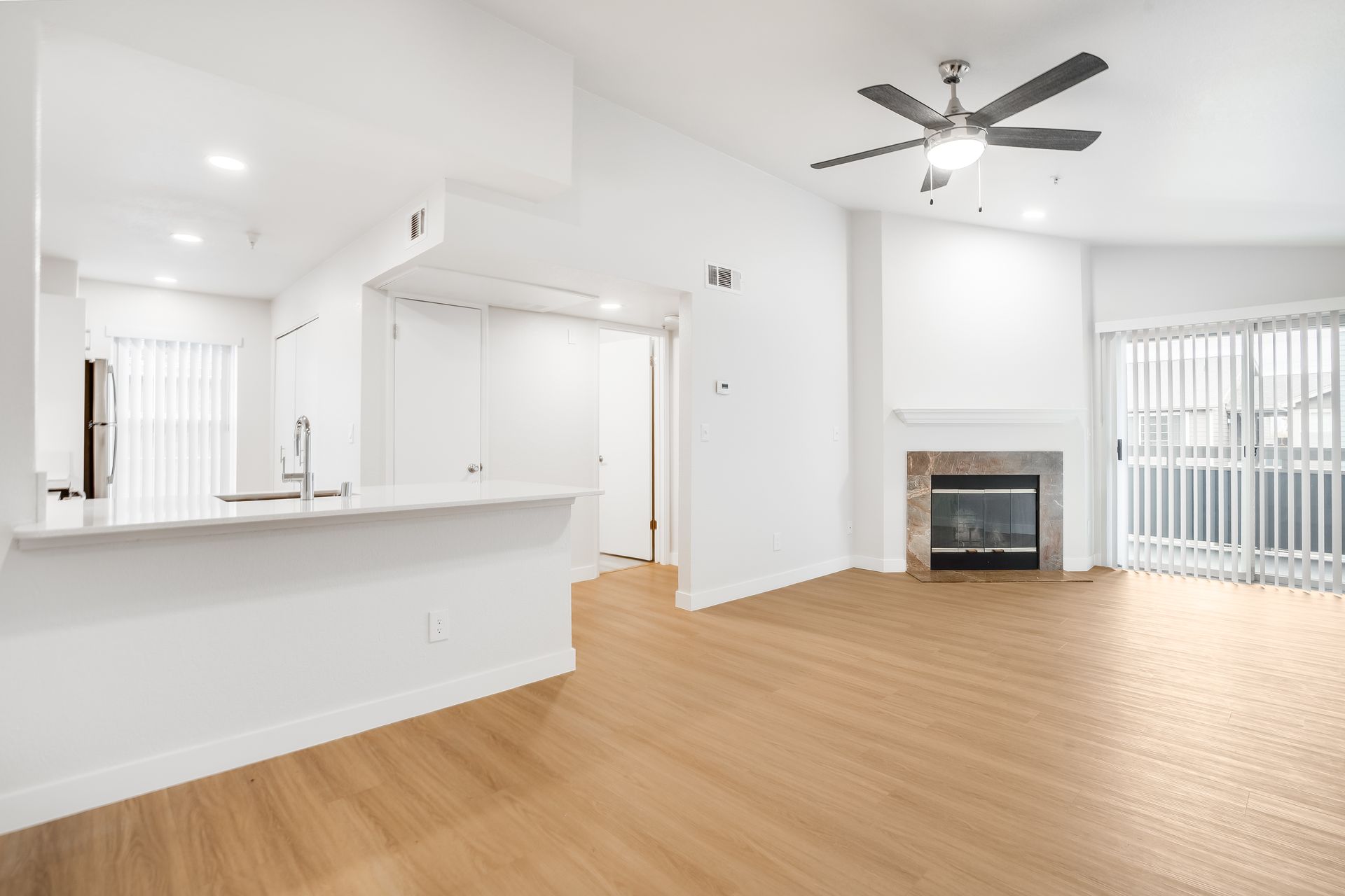 Bright empty living room with hardwood floors, white walls, ceiling fan, fireplace, and open kitchen counter