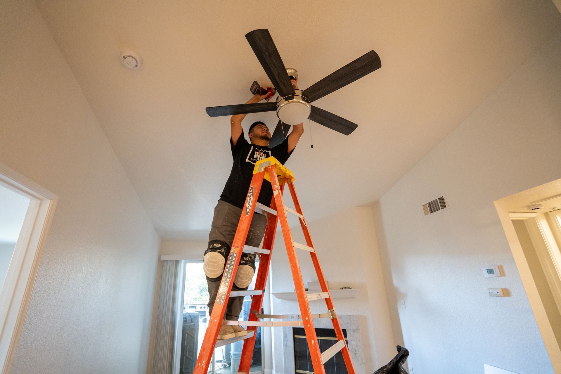 Person on orange ladder installing a ceiling fan in a bright room.
