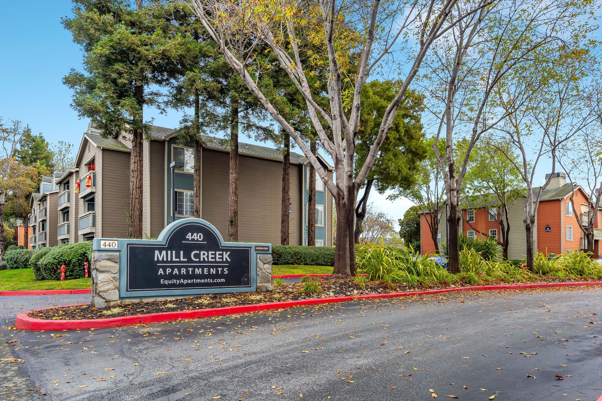 Mill Creek apartment complex sign at a tree-lined entrance with buildings and parked cars in the background