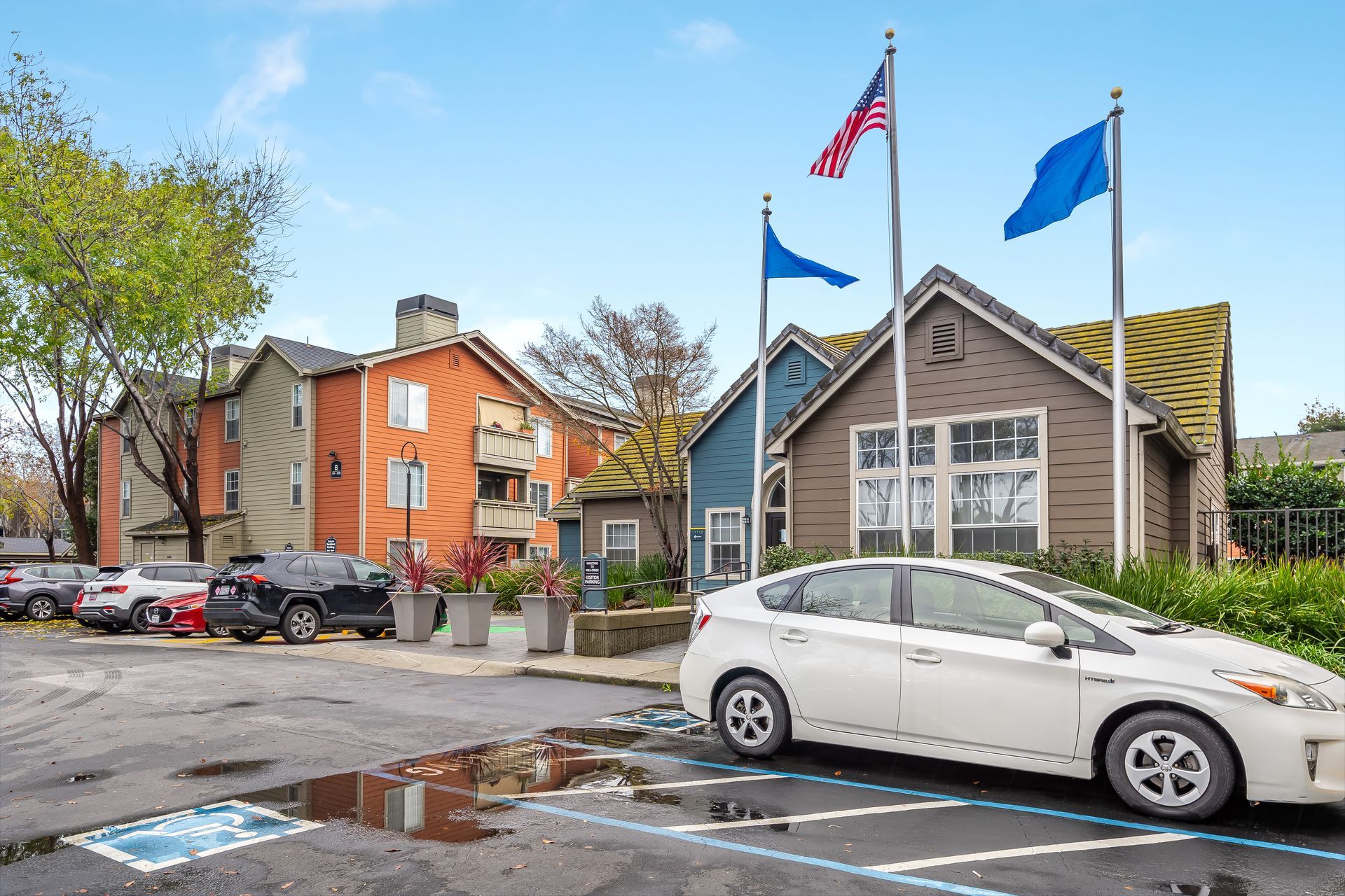 Parking lot with white car, storefronts, and three flags under a blue sky