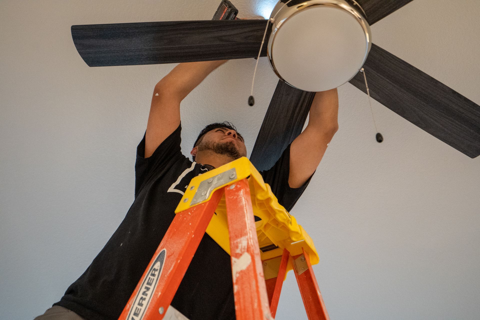 Person on orange ladder installing a ceiling fan with a yellow tool