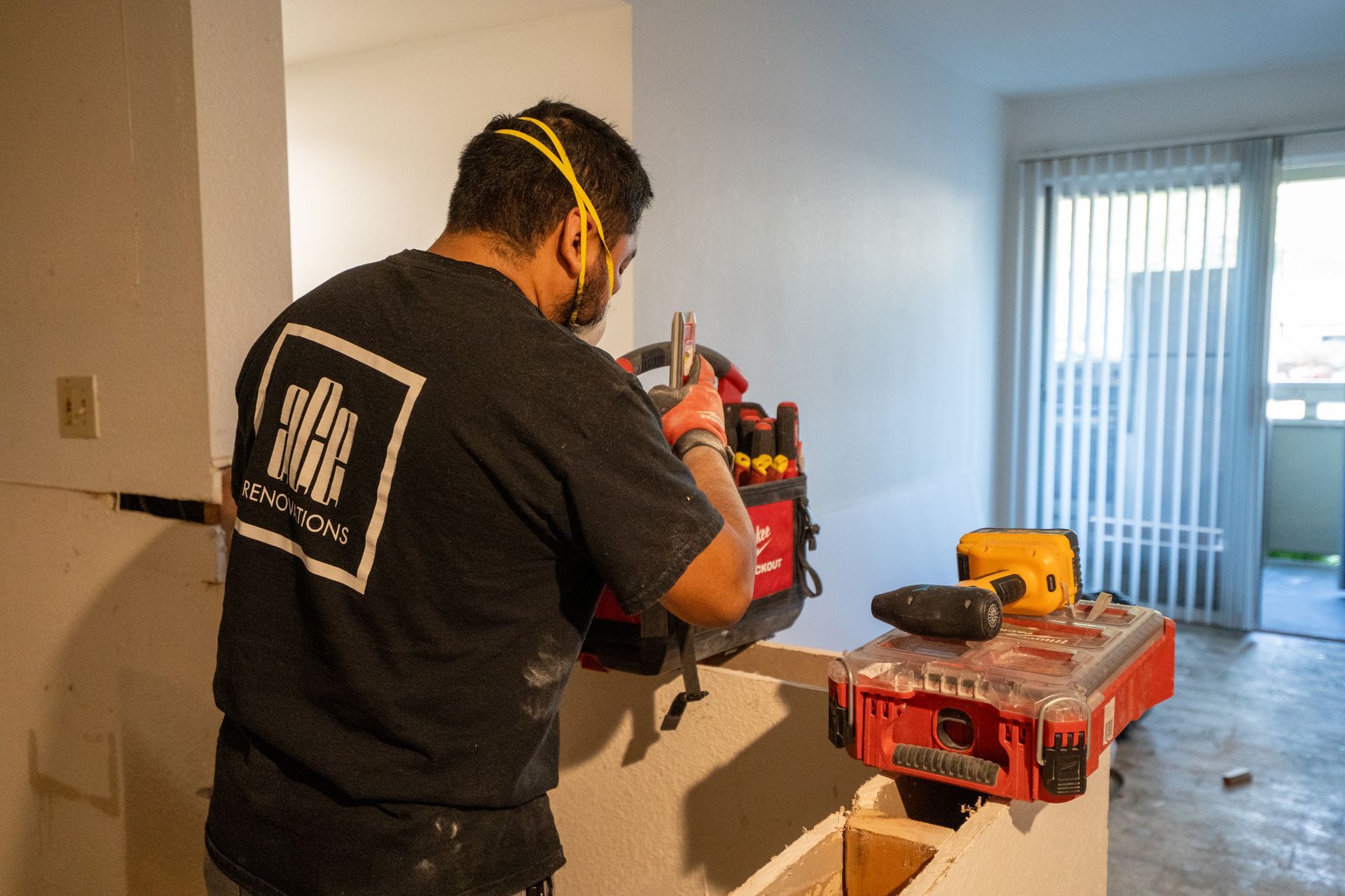 Worker sanding a red wall in a bright room, with tools on a nearby workbench.