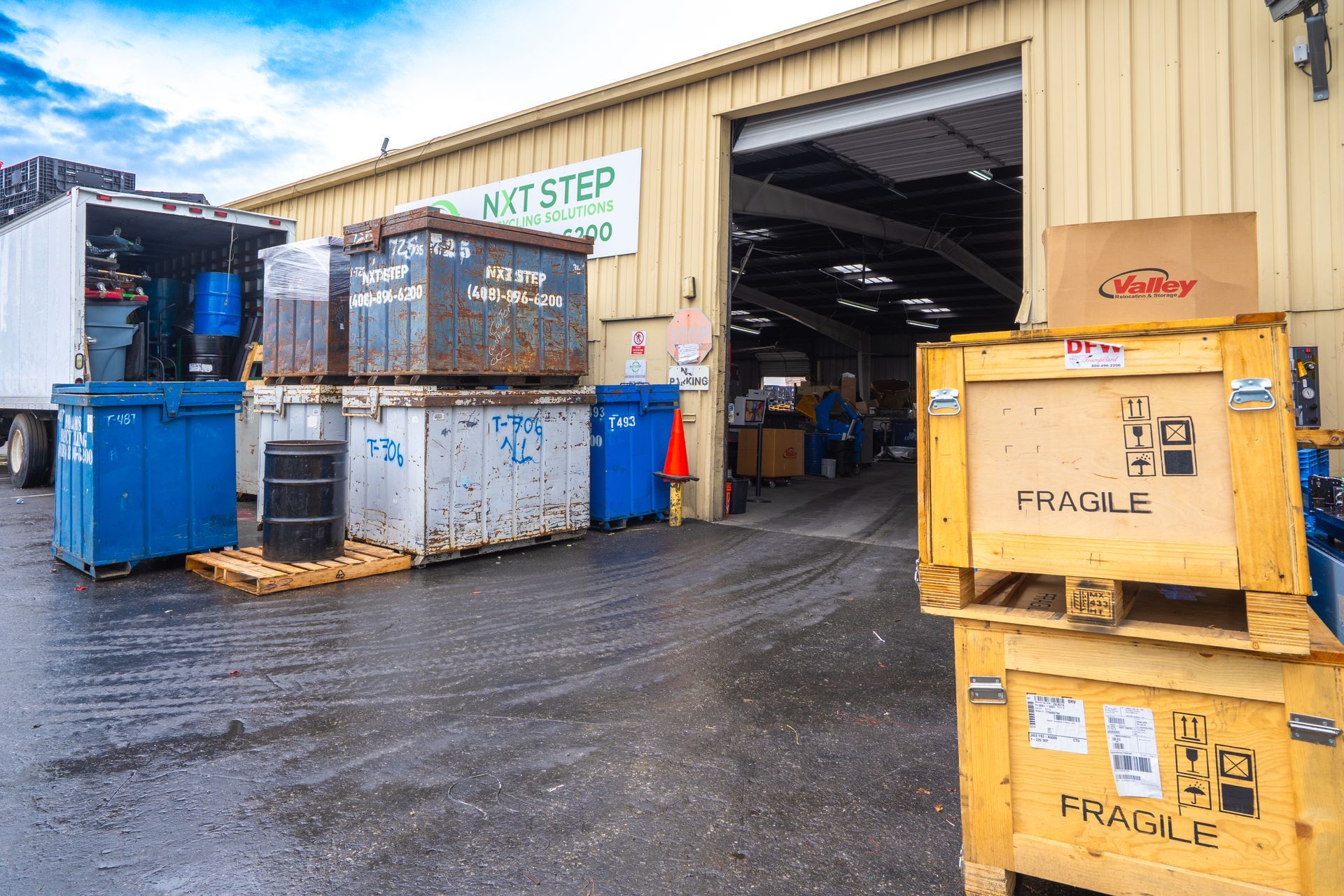 Exterior of warehouse with shipping crates, dumpsters, and a semi-truck. Wet pavement reflects the cloudy sky.