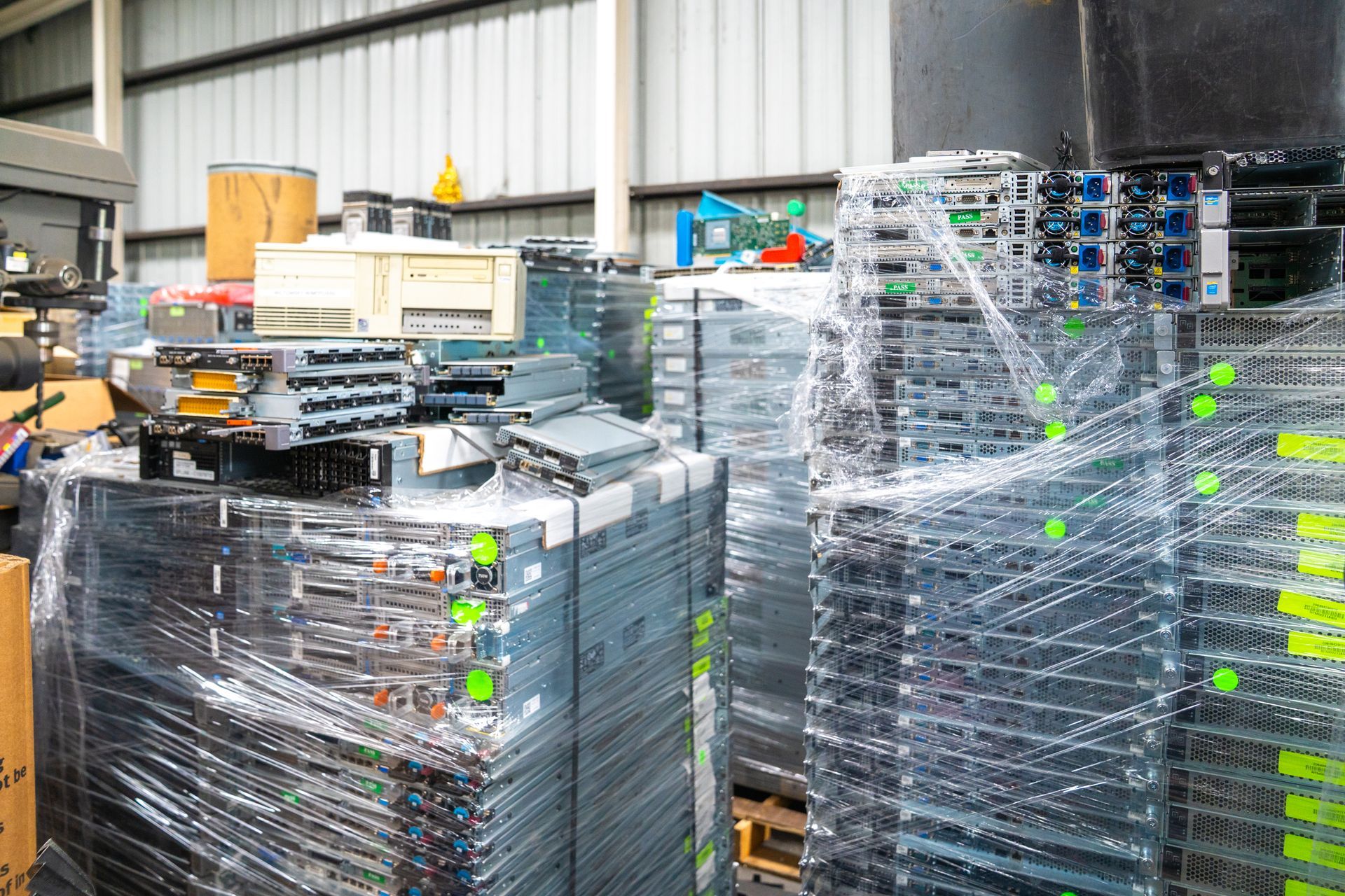 Piles of bundled computer server racks on pallets in a warehouse, wrapped in plastic.