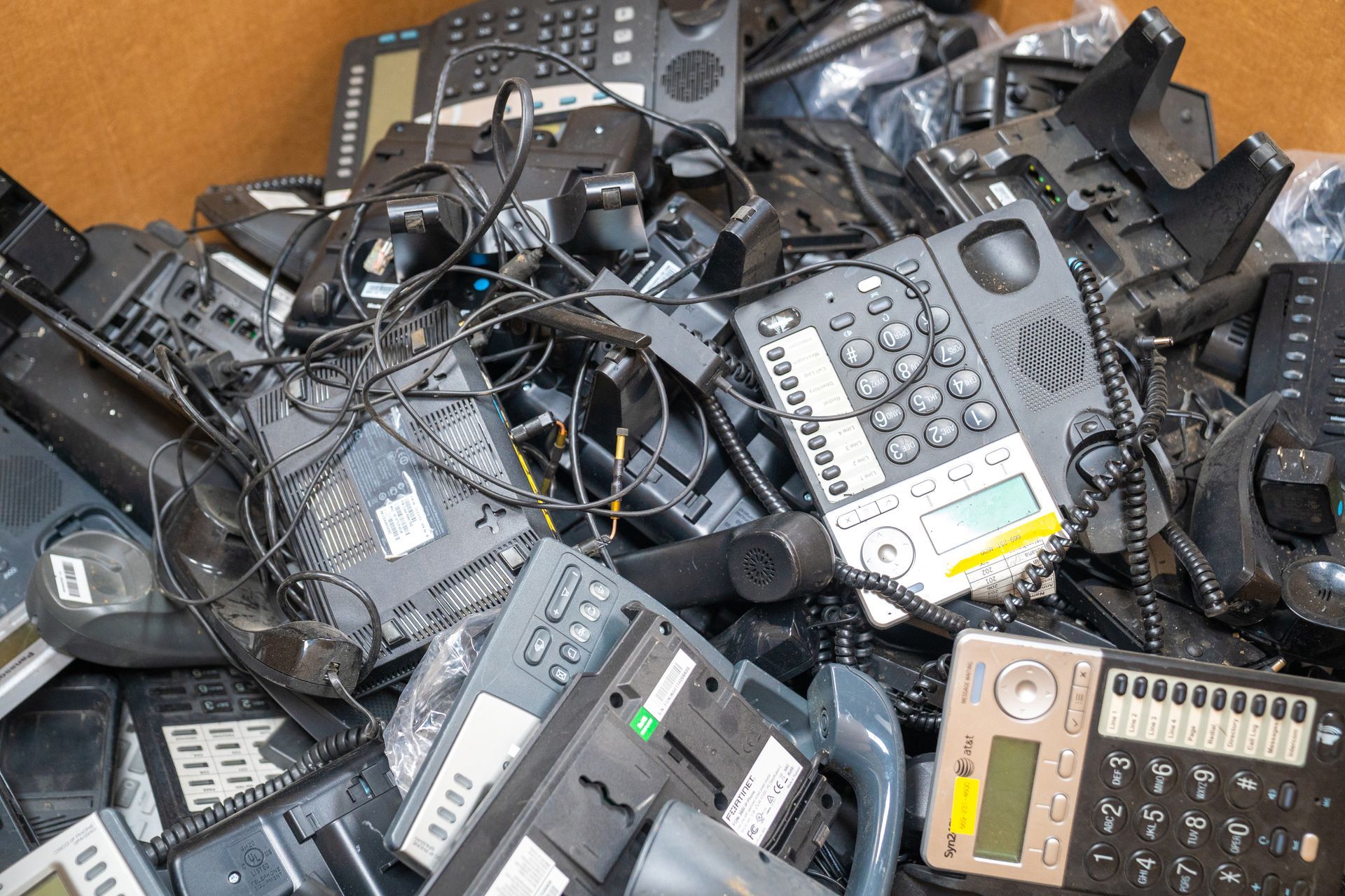 Pile of black business telephones in a box, tangled with wires and chargers.