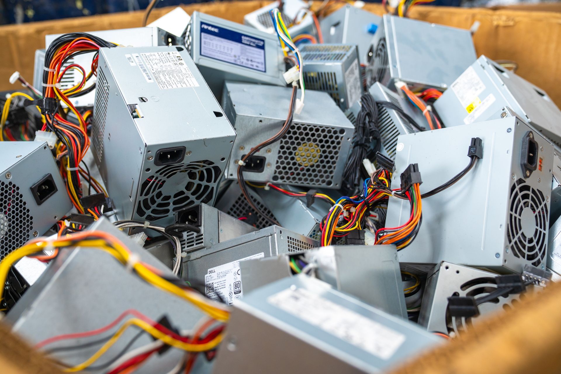 Pile of silver computer power supply units with colorful wires in a cardboard box.