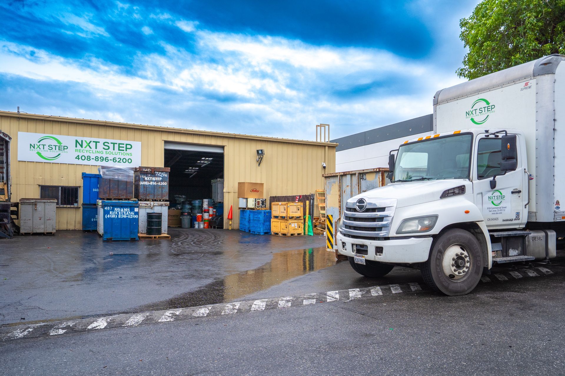 White truck parked in front of a recycling center with blue bins and a yellow building under a cloudy sky.
