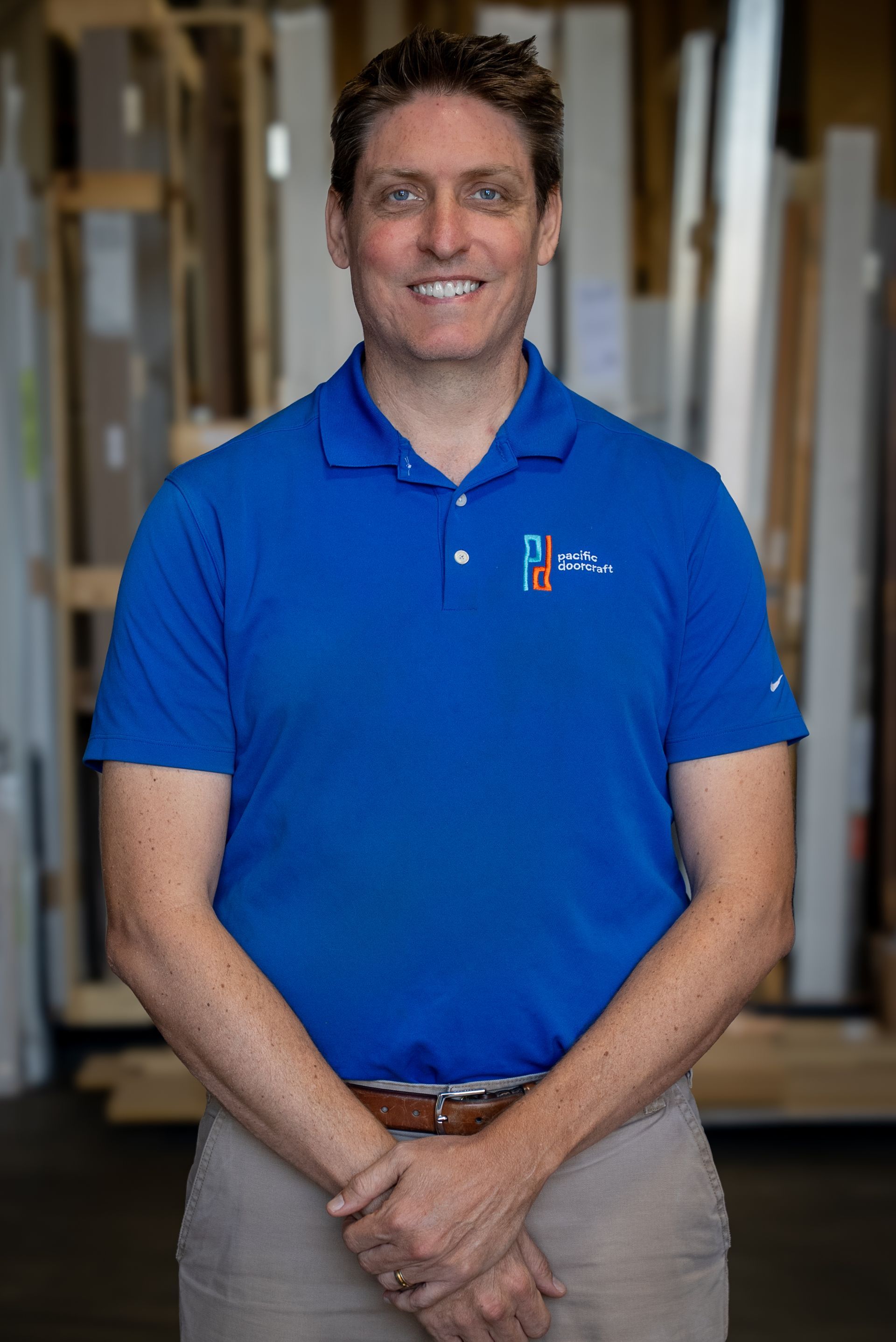 Man standing indoors in a blue polo shirt, smiling with hands clasped in front of him.