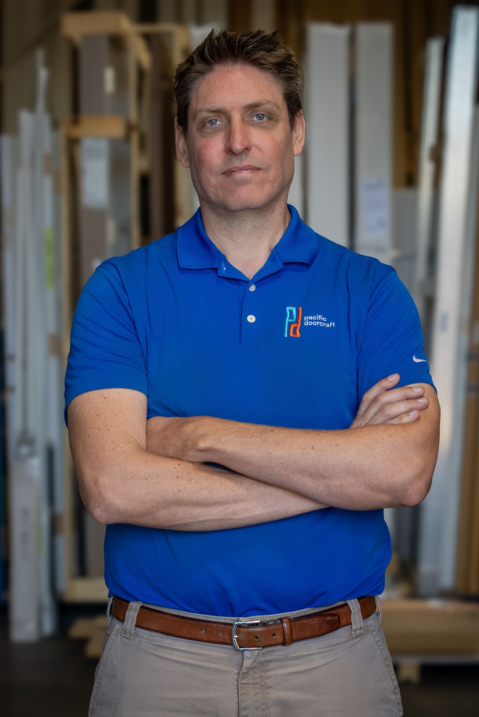 Man standing with arms crossed in a blue polo shirt in a workshop or warehouse setting