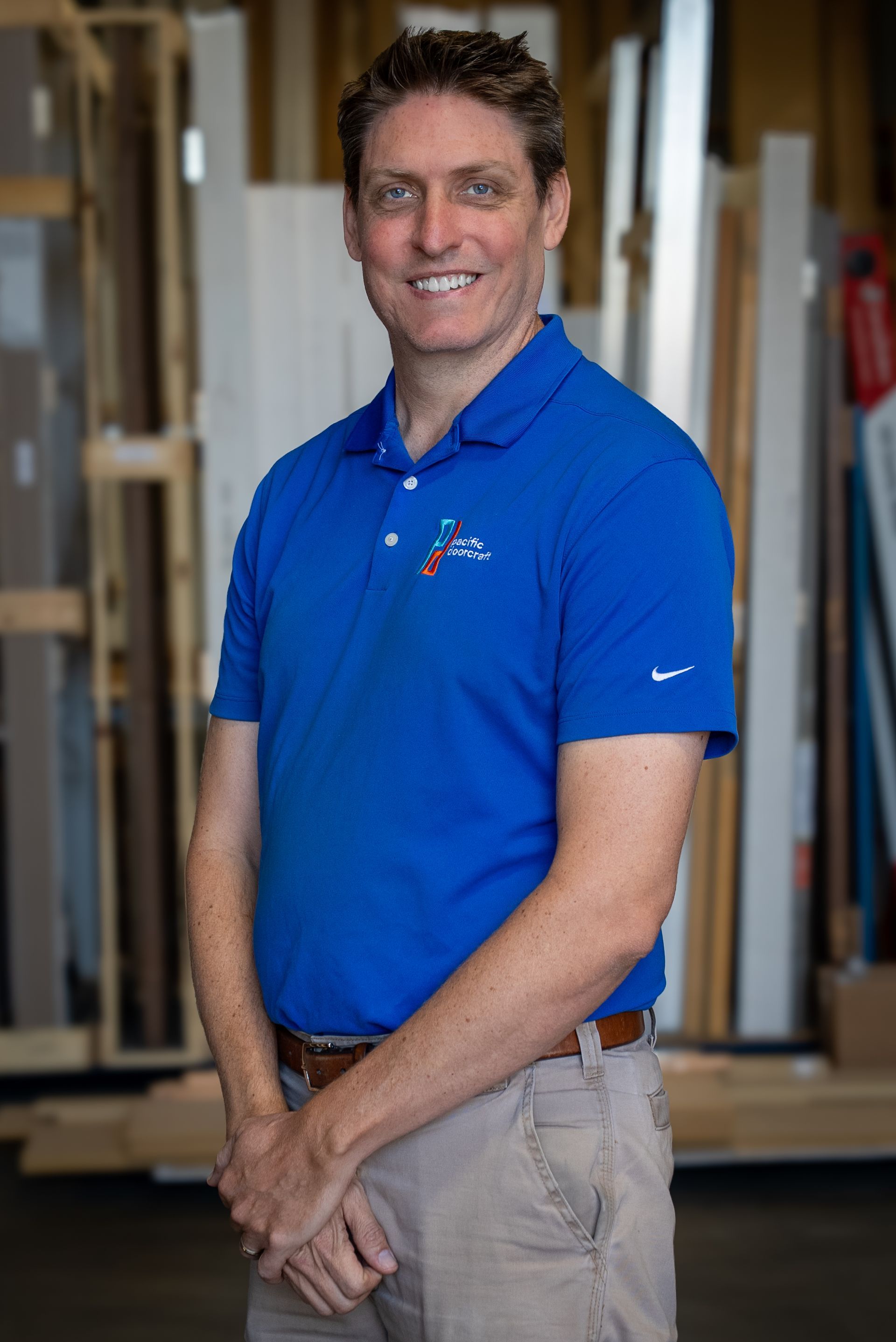 Man in a blue polo shirt standing indoors in a workshop, smiling with hands clasped.