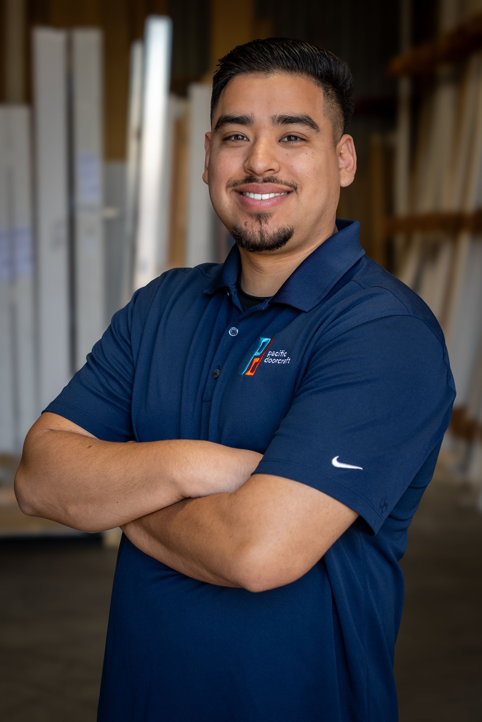 Man standing with arms crossed, wearing a navy polo, in an office or workshop setting.