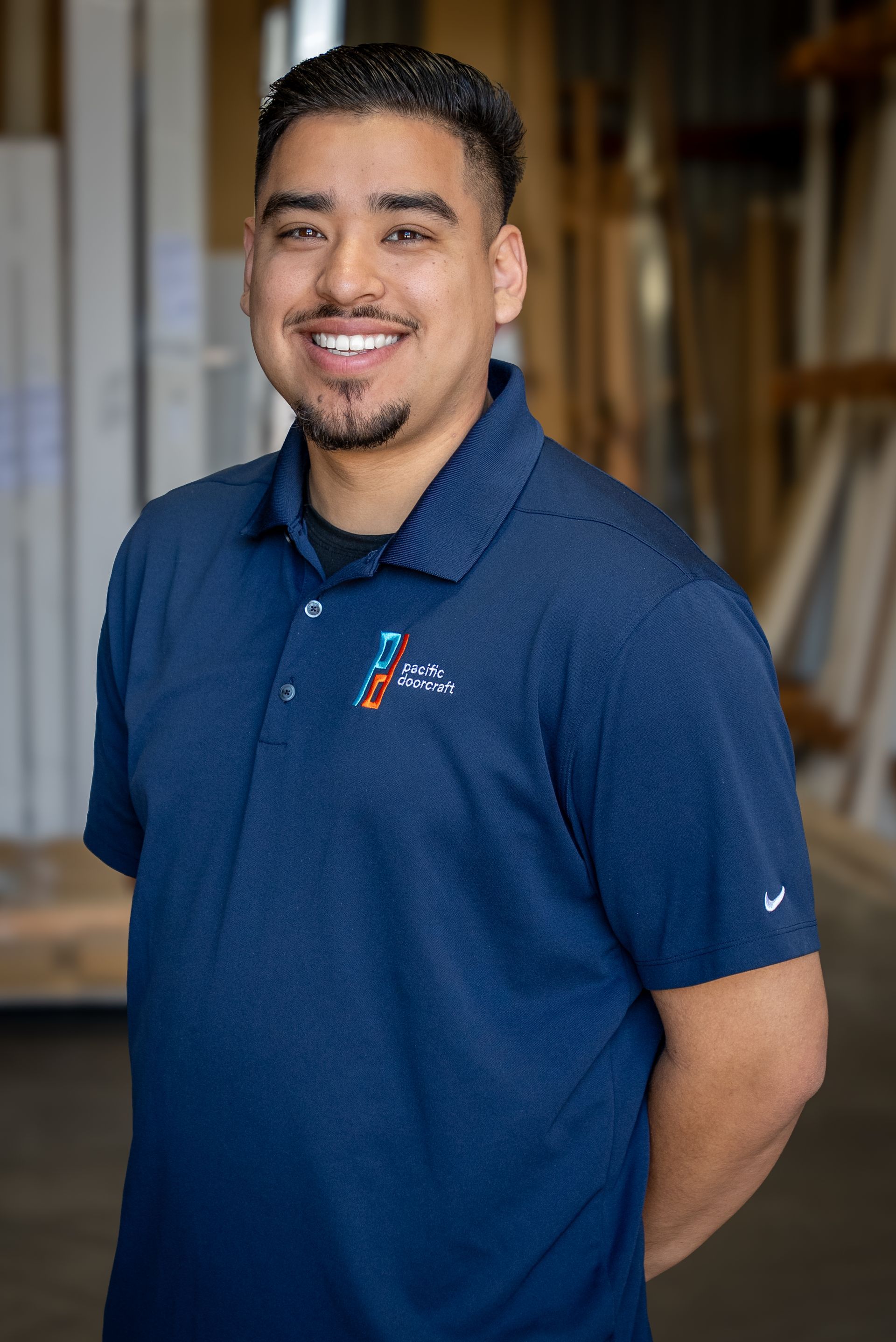Smiling man in a navy polo shirt standing with hands behind his back indoors.