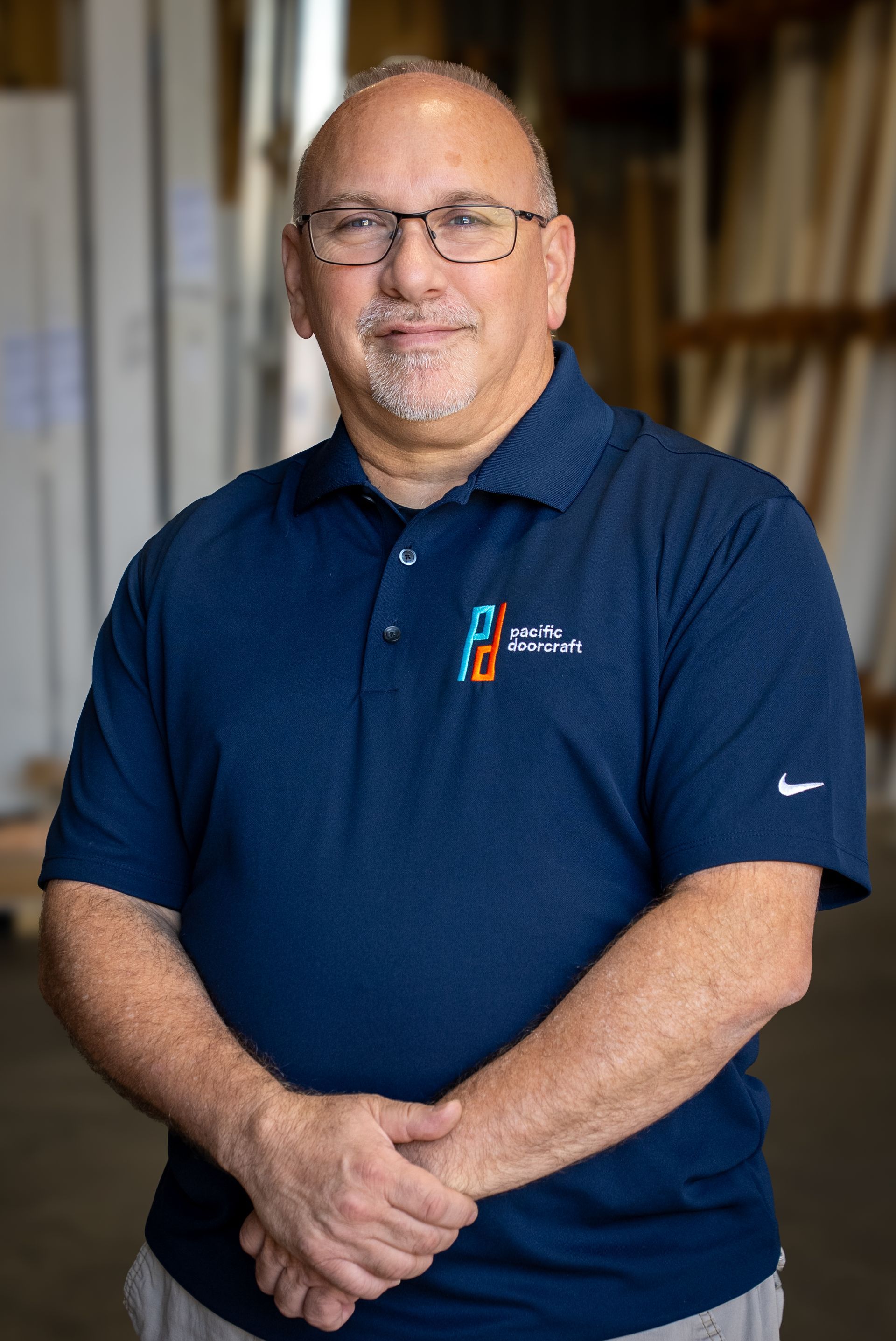 Smiling man in navy polo shirt standing indoors with hands clasped, bookshelves in the background