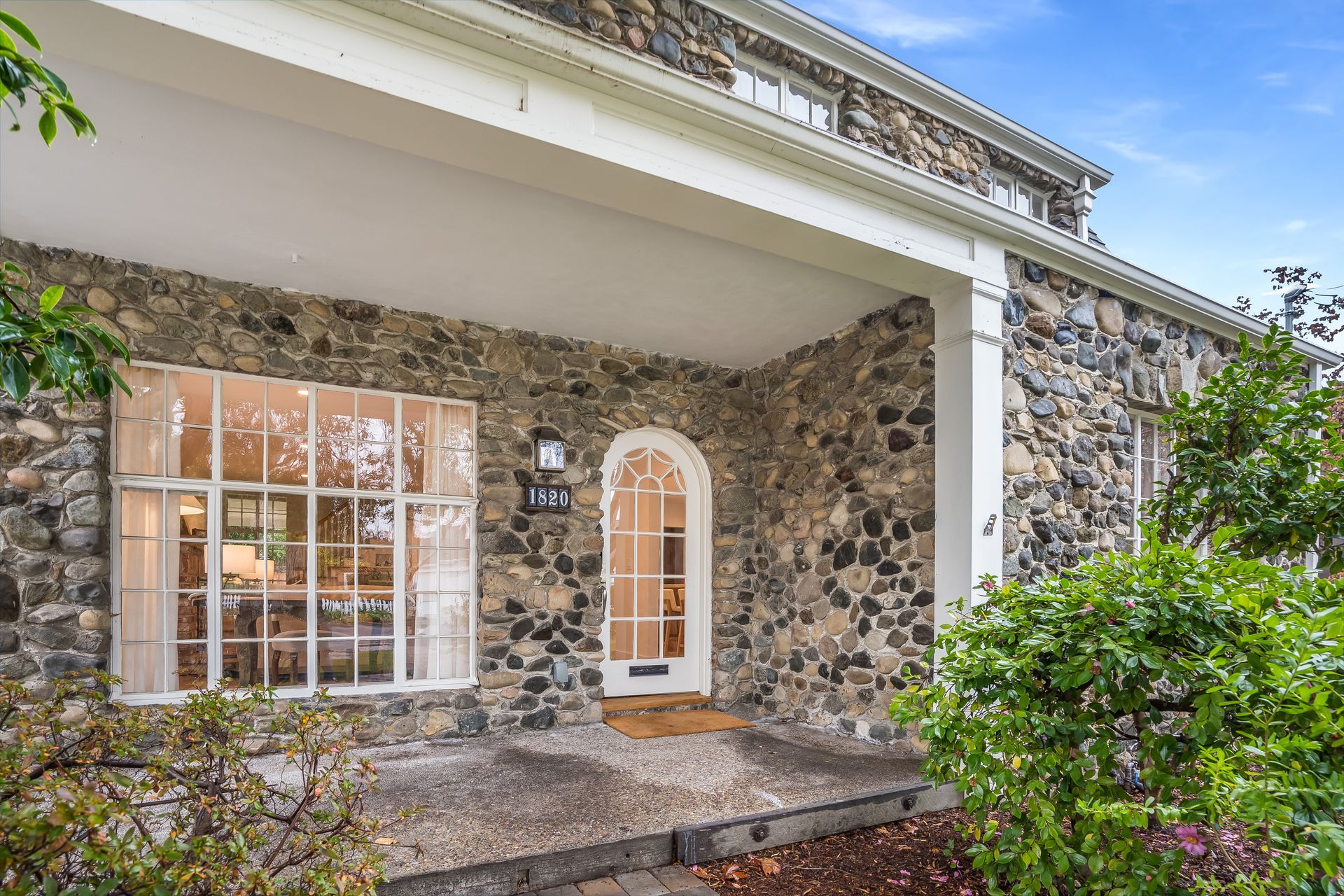 Stone house porch with white columns, arched front door, and large window surrounded by greenery