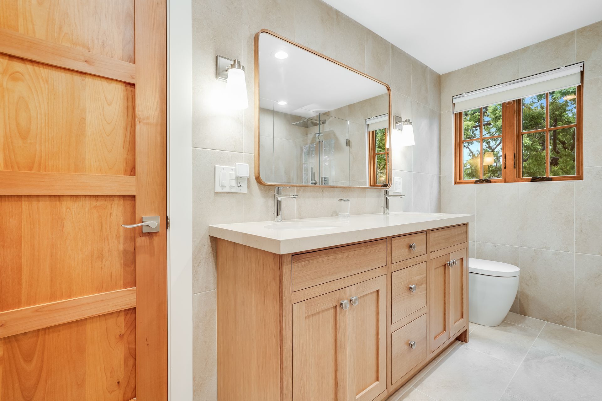 Bright bathroom with wood vanity, large mirror, toilet, and window by the shower area