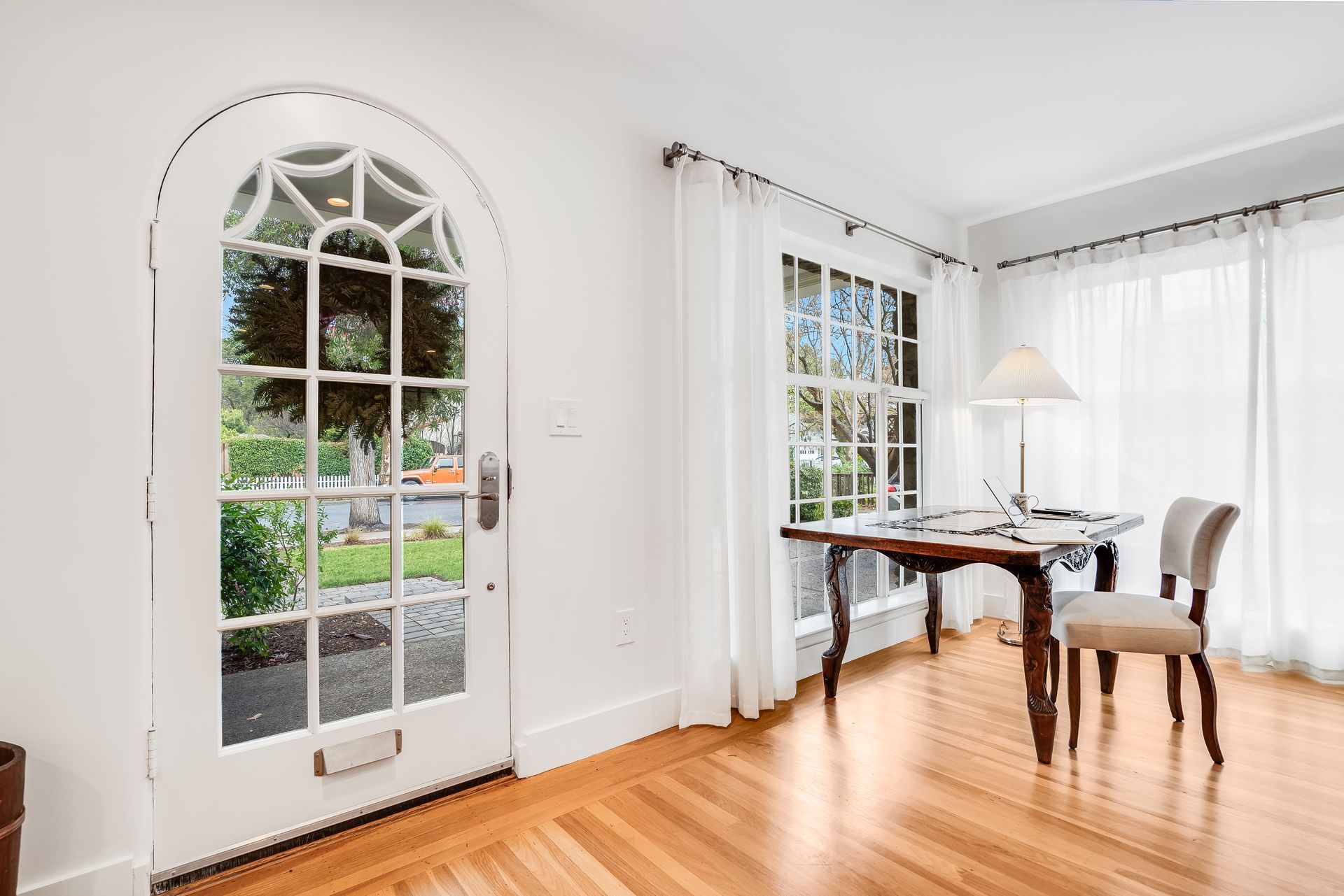 Bright dining area with arched glass door, white curtains, wooden floor, table, chairs, and floor lamp.
