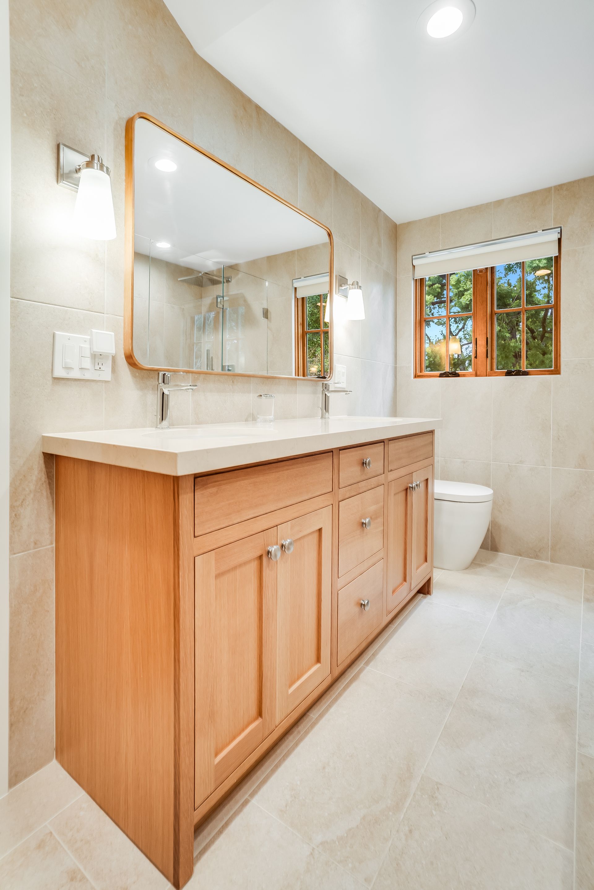 Bright bathroom with light wood vanity, large mirror, and window beside a toilet