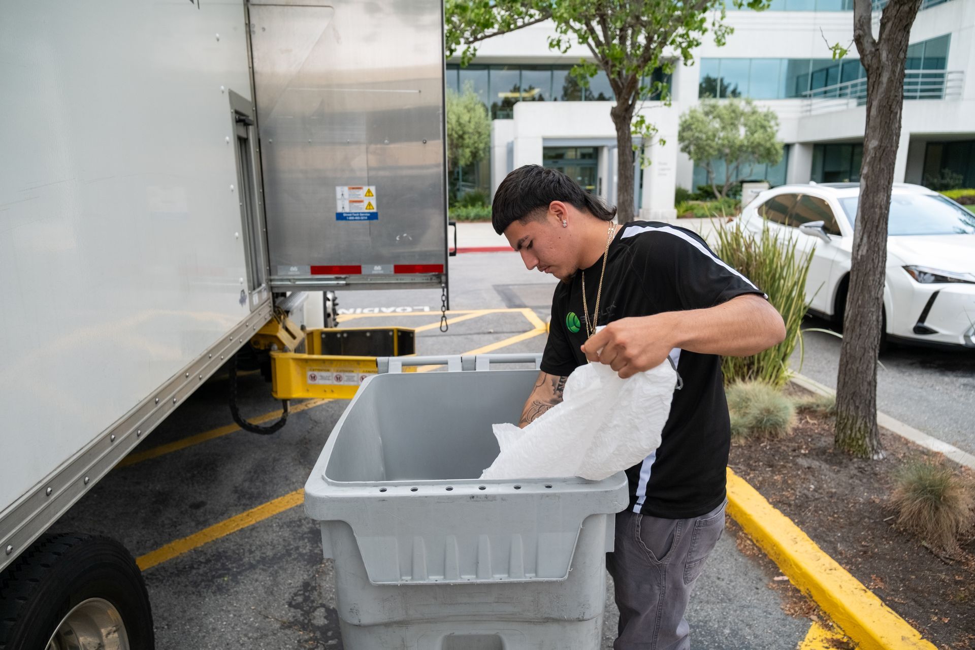 Man putting items from a truck into a gray bin outdoors.  Truck is parked, with a modern building in the background.