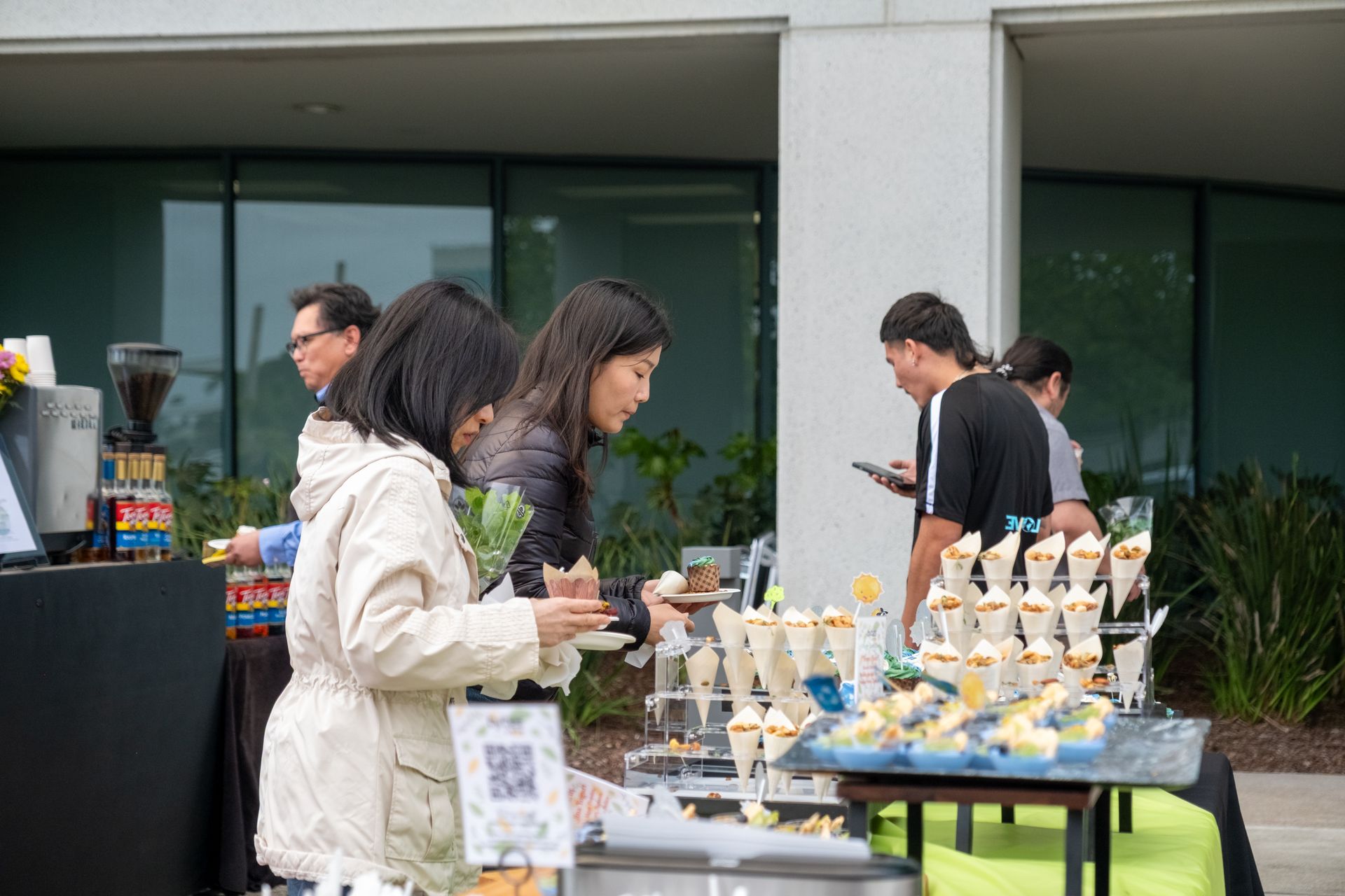 People at an outdoor food table with various small appetizers and drinks.