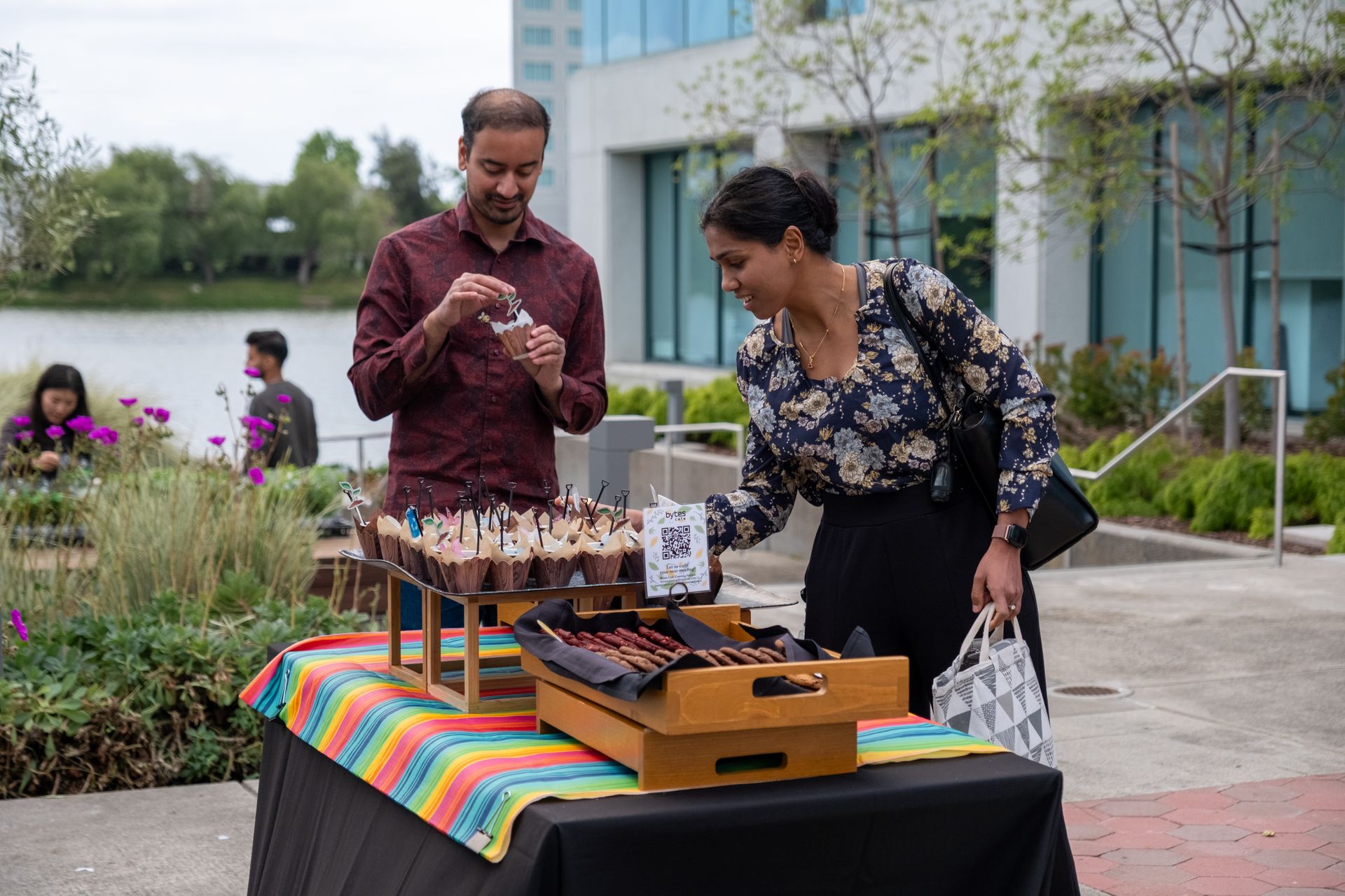 People at an outdoor market examining cupcakes and treats; lake and building background.