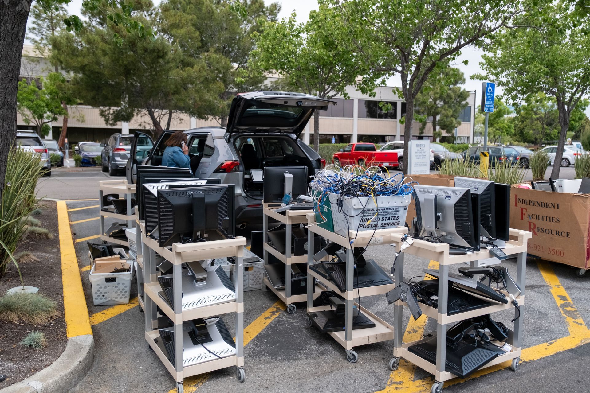 Computers and monitors on wheeled carts near a car in a parking lot, possibly for recycling or disposal.