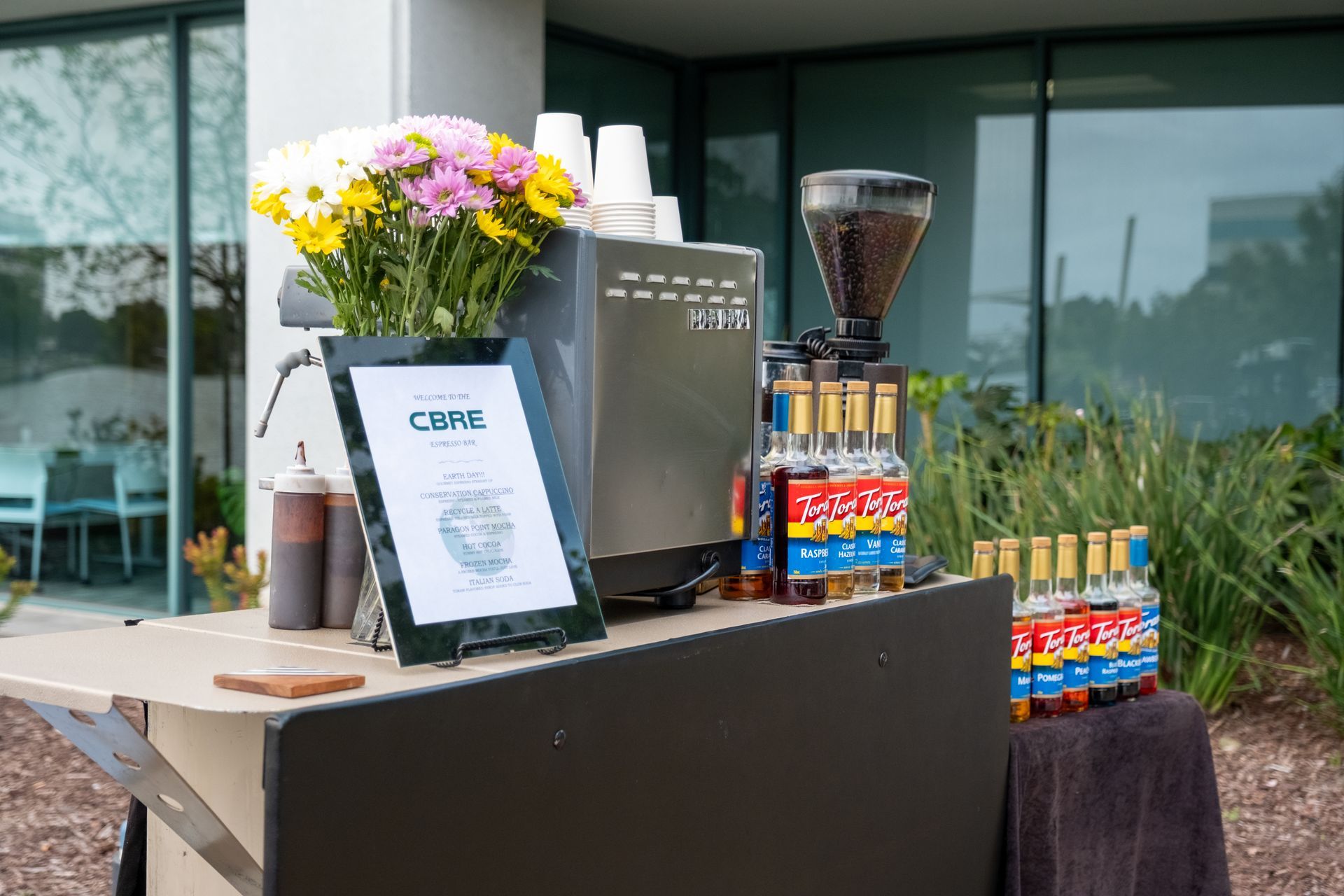 Coffee cart with espresso machine, syrups, cups, and flowers outside a building.