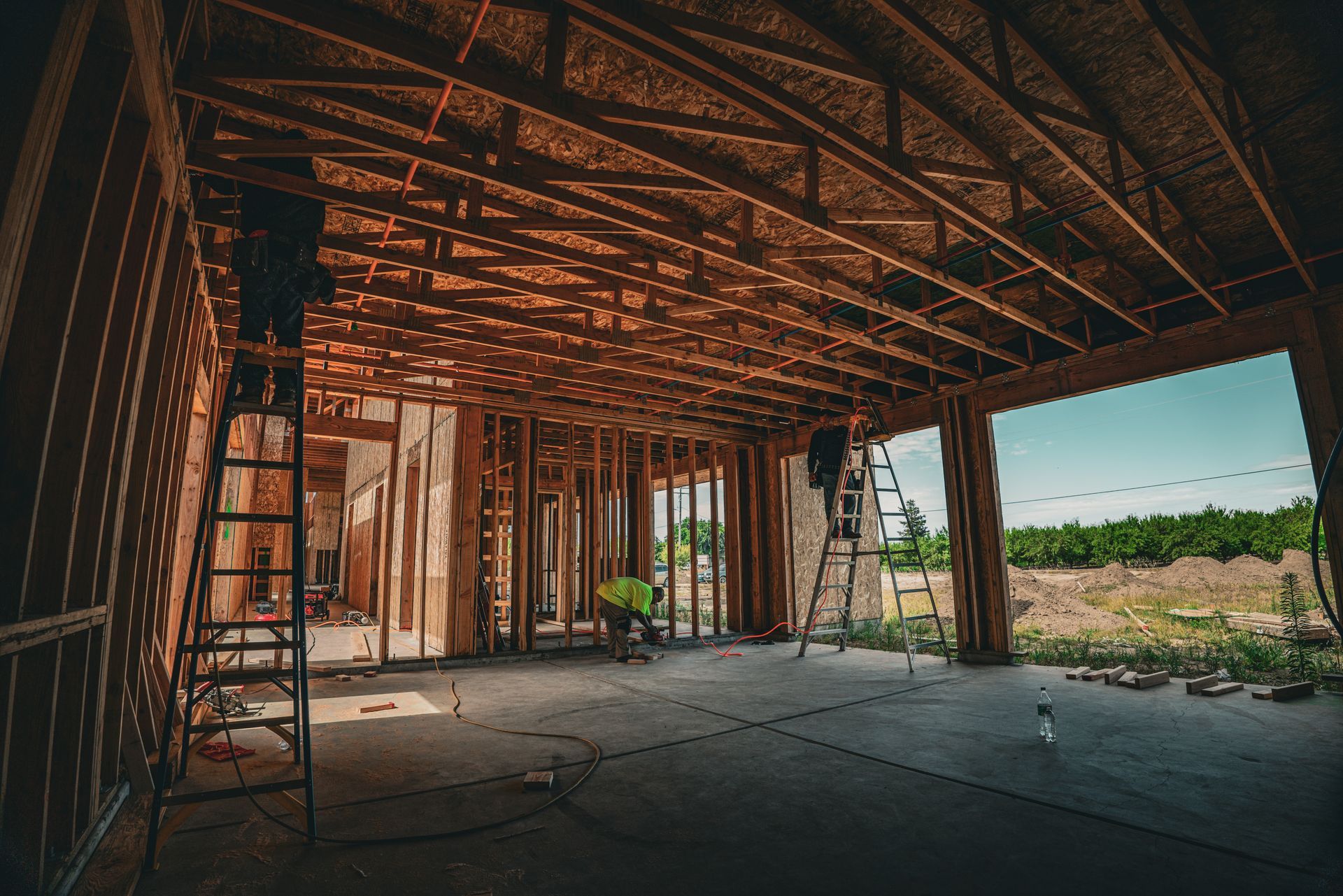 Interior of unfinished house framing with exposed beams, concrete floor, and open view to grassy field