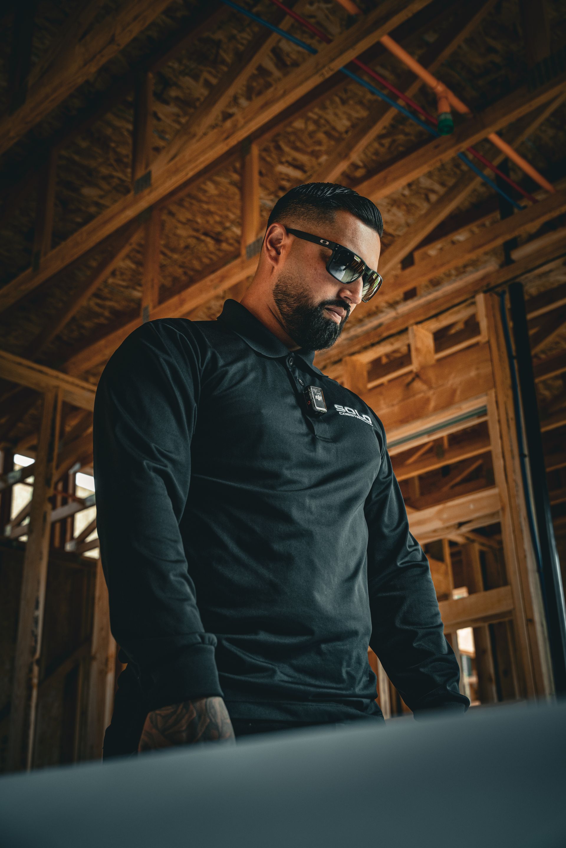 Man in black shirt and sunglasses standing in a wooden-framed construction site, looking downward