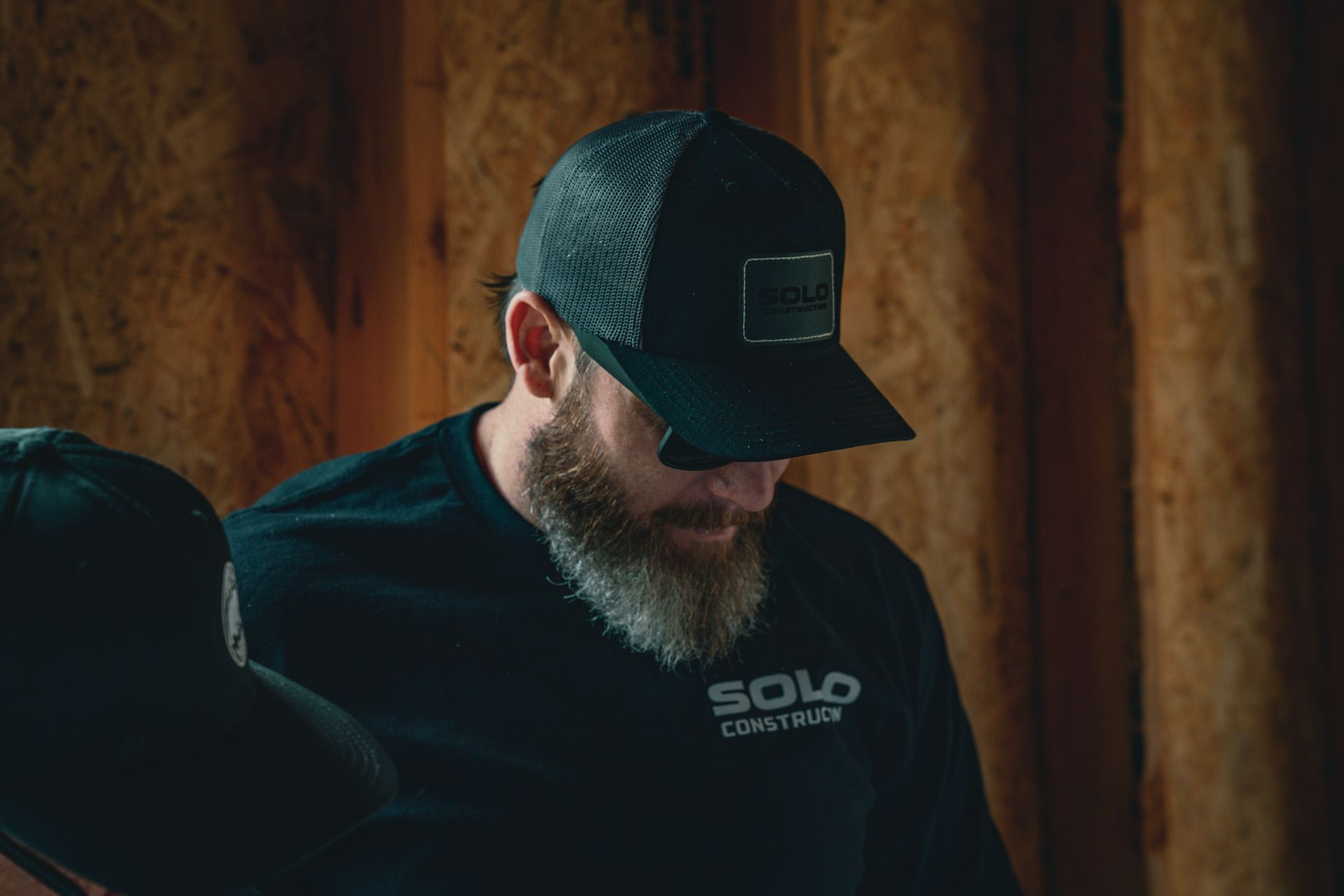 Bearded man in a dark cap and shirt, looking down in a warm-lit wooden room