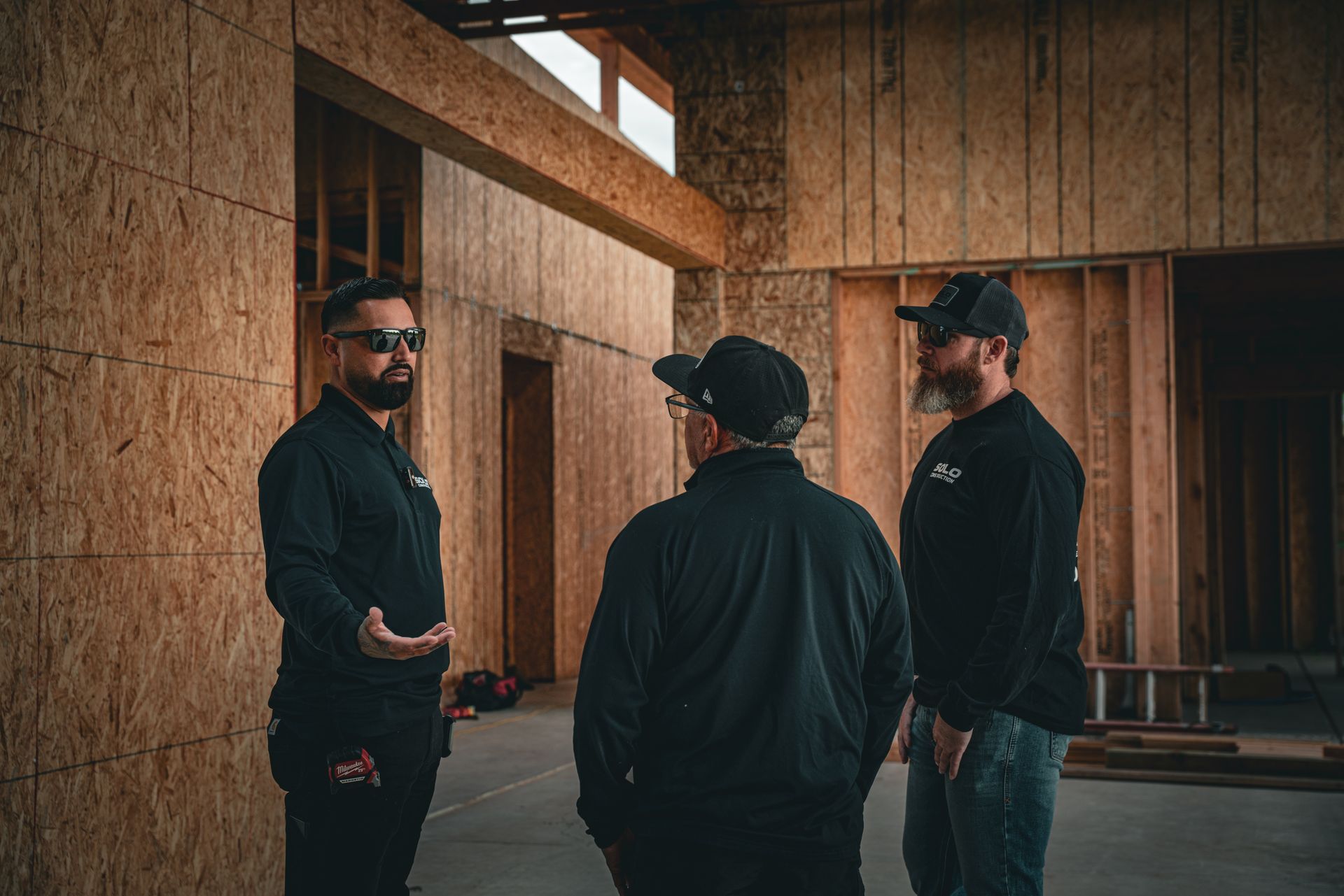 Three men in dark clothing stand talking inside an unfinished wooden building.