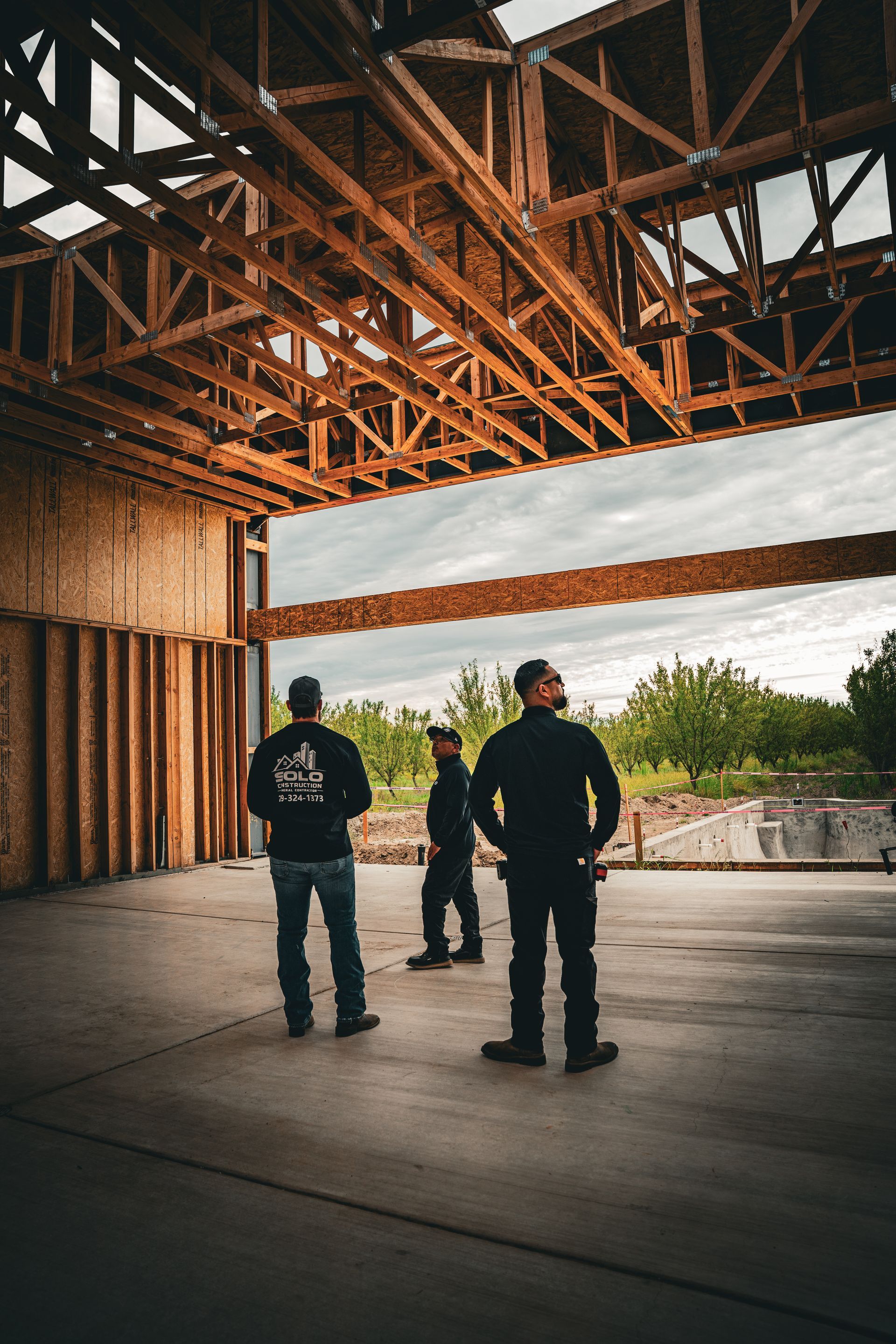 Two people stand inside an unfinished building with exposed wooden beams and a view of trees outside.