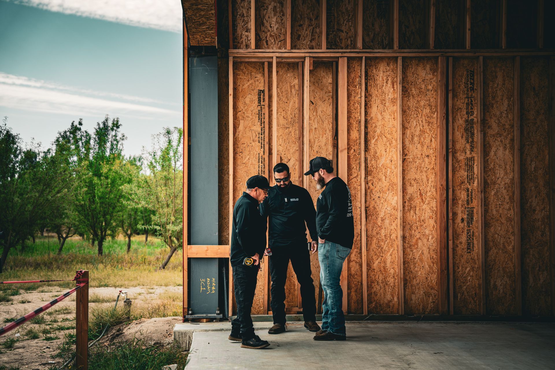 Three people stand beside a wooden wall in an unfinished building opening onto a grassy field.