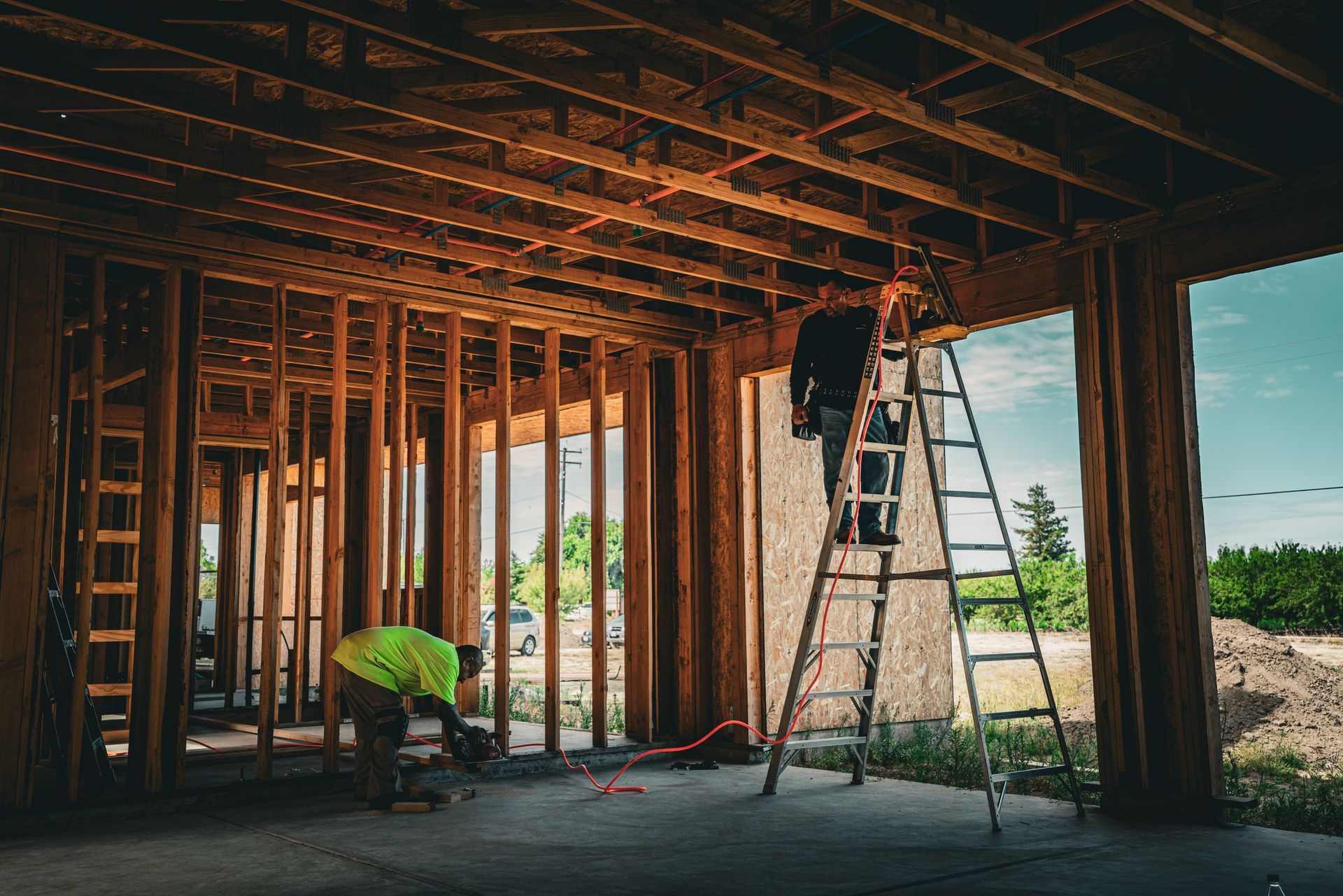 Construction workers framing a house interior with exposed wooden studs, ladders, and tools under a partially finished roof