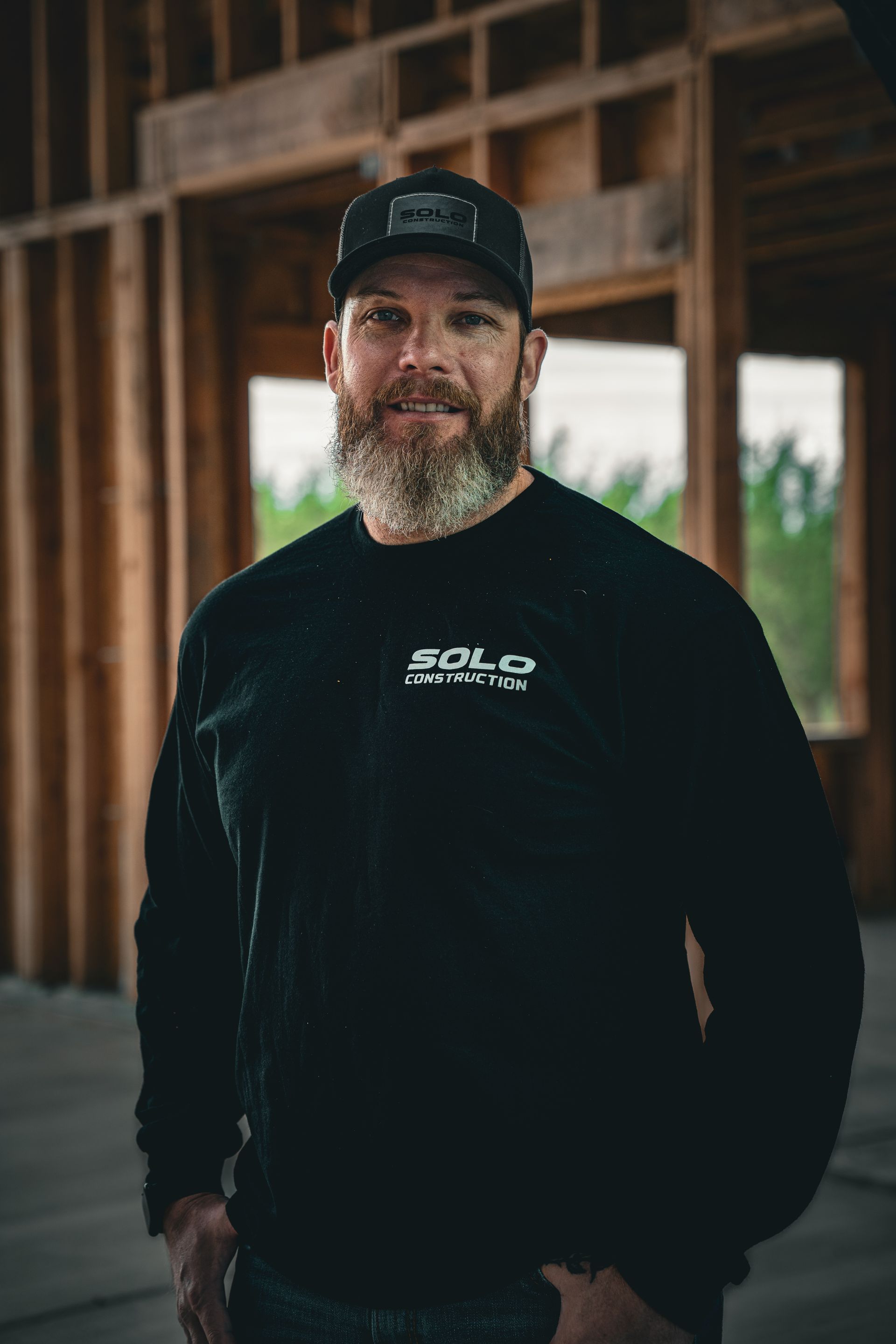 Smiling bearded man in black cap and sweatshirt standing in a wooden-framed building.