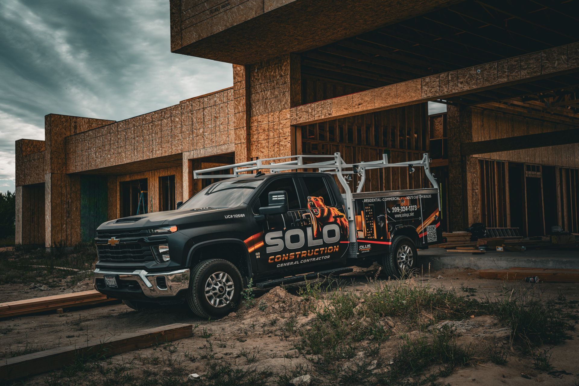 Black off-road pickup with roof rack parked by a modern building in a rainy, overcast setting