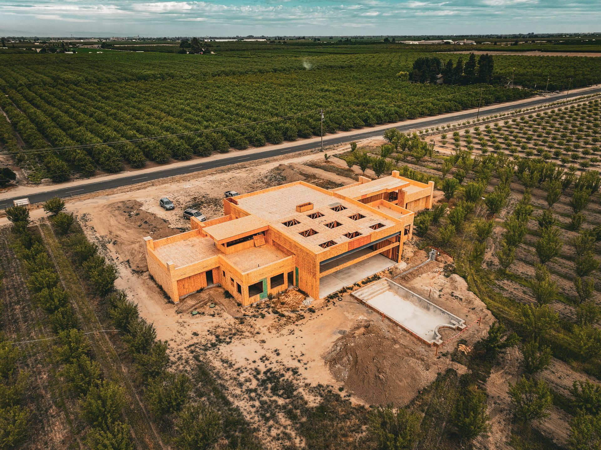 Aerial view of an orange building under construction beside a dirt road, surrounded by green fields.